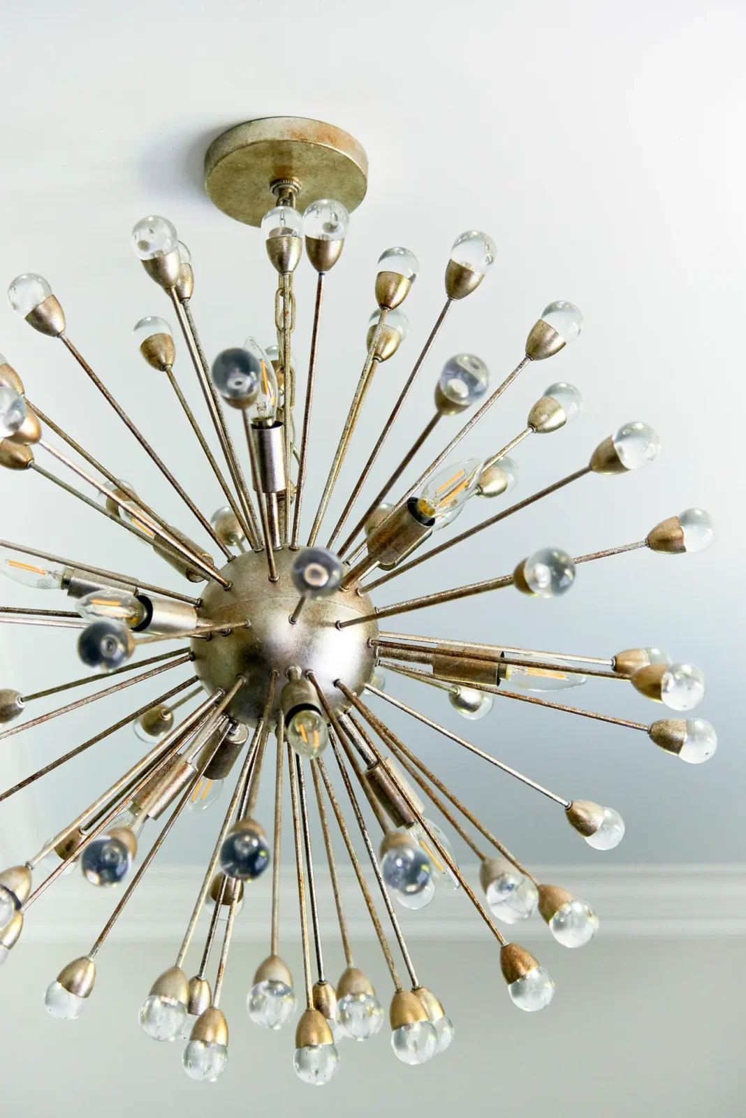 A decorative, antique gold sputnik chandelier hangs from a white ceiling, featuring radiating arms tipped with clear glass teardrop bulbs.