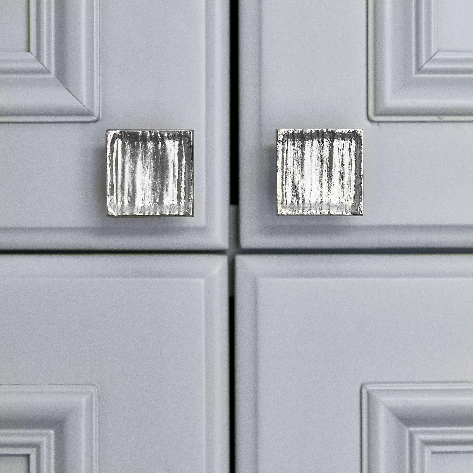 A close-up of light gray shaker style cabinet doors featuring two square, metallic knobs with a vertical textured surface.