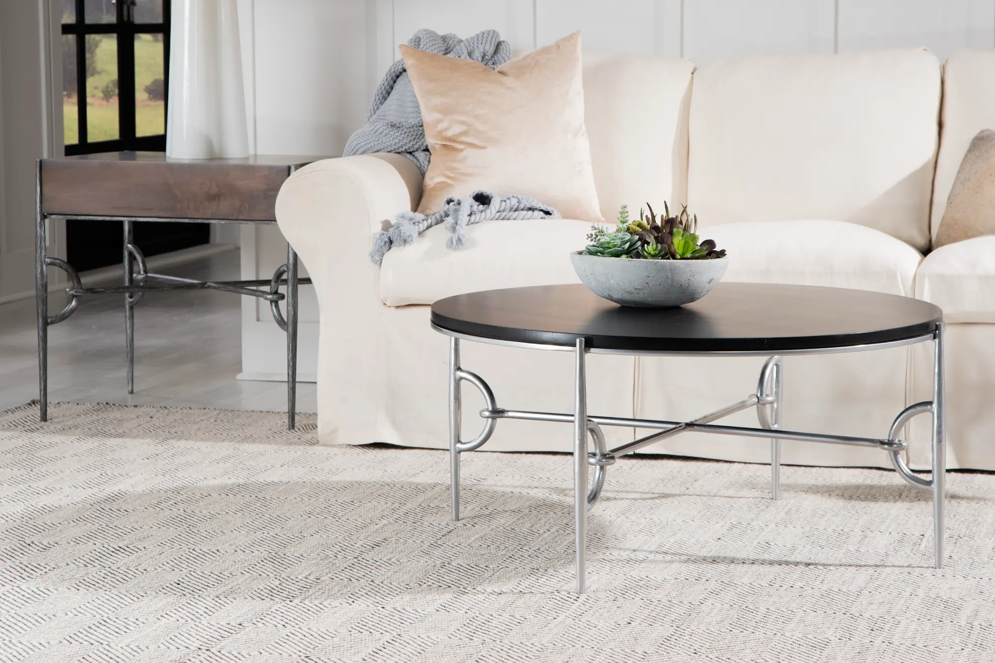 A sophisticated living room features The Paddock Collection's black round coffee table and wood side table, all supported by intricate silver metal bases.
