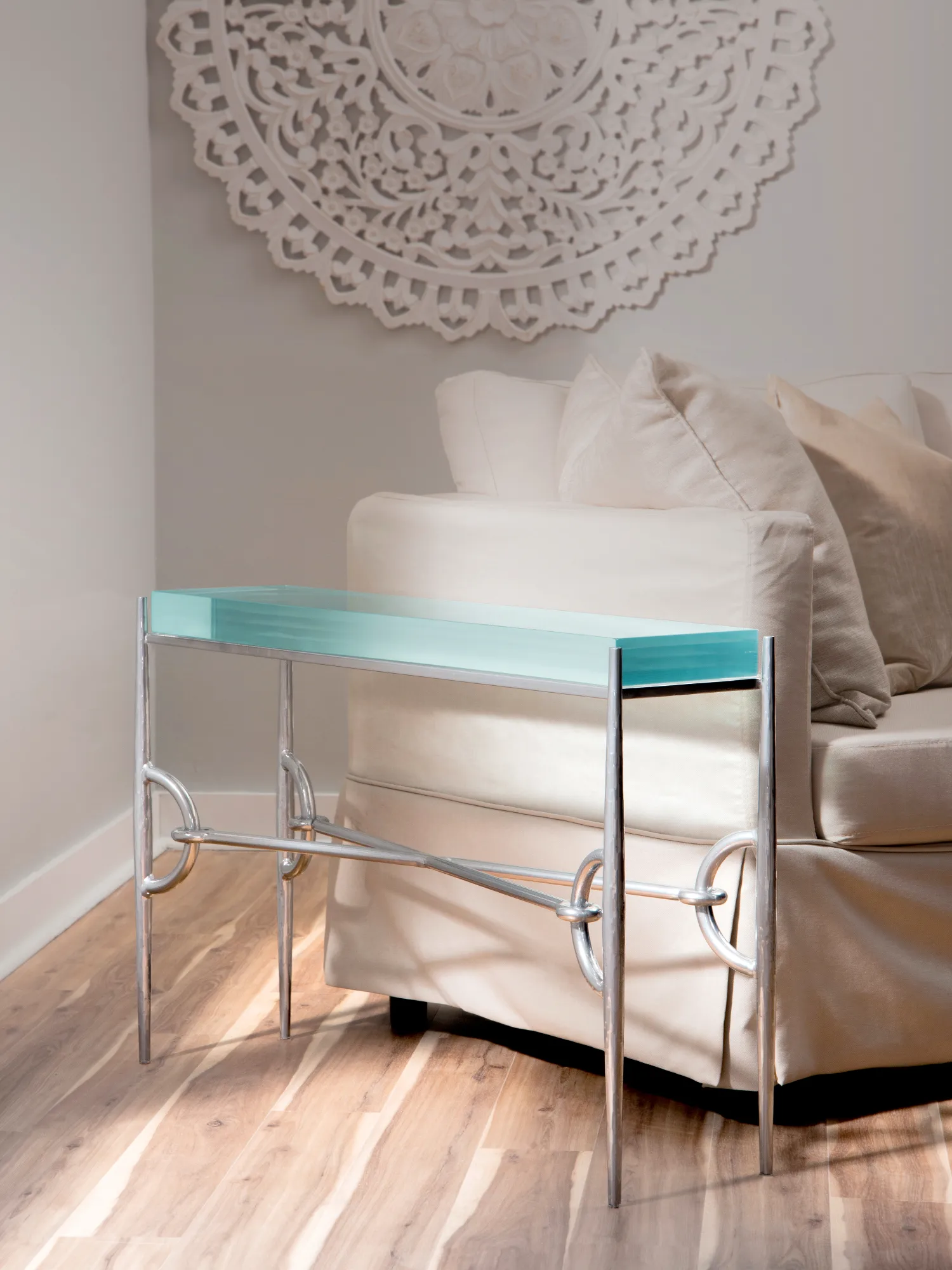 The Paddock Collection chairside console table with a thick aqua glass top and polished chrome metal base sits next to a light-colored sofa beneath an ornate white wall medallion.
