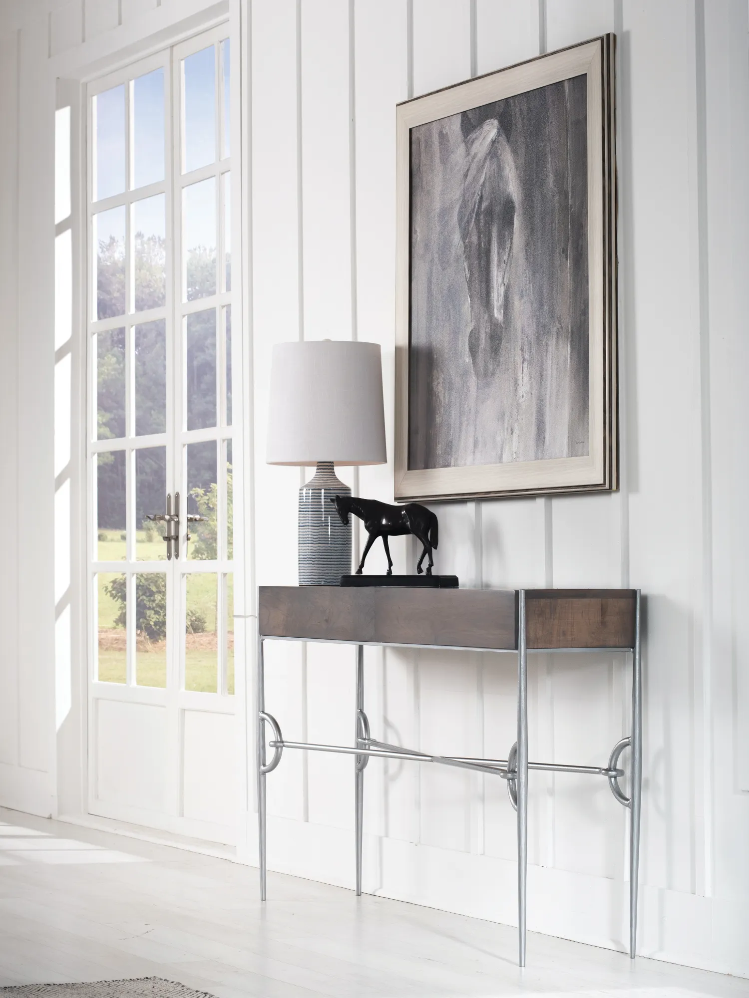 The Paddock Collection console table with wooden top and chrome equestrian accents holds a lamp and horse sculpture against bright white walls, next to a large window.