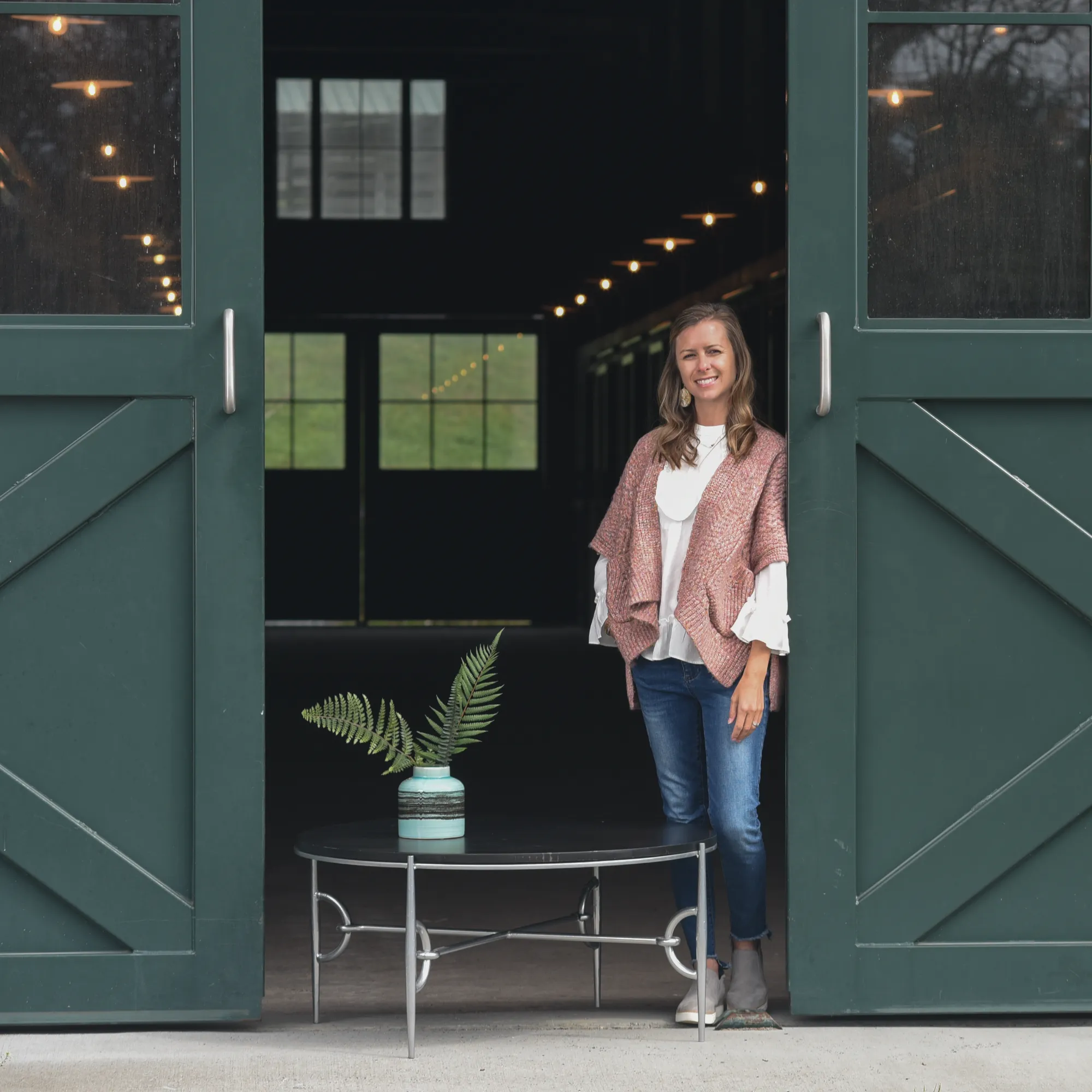 Interior designer Lauren Clement smiles, standing in a barn doorway next to The Paddock Collection round coffee table styled with a teal vase and fern leaves.