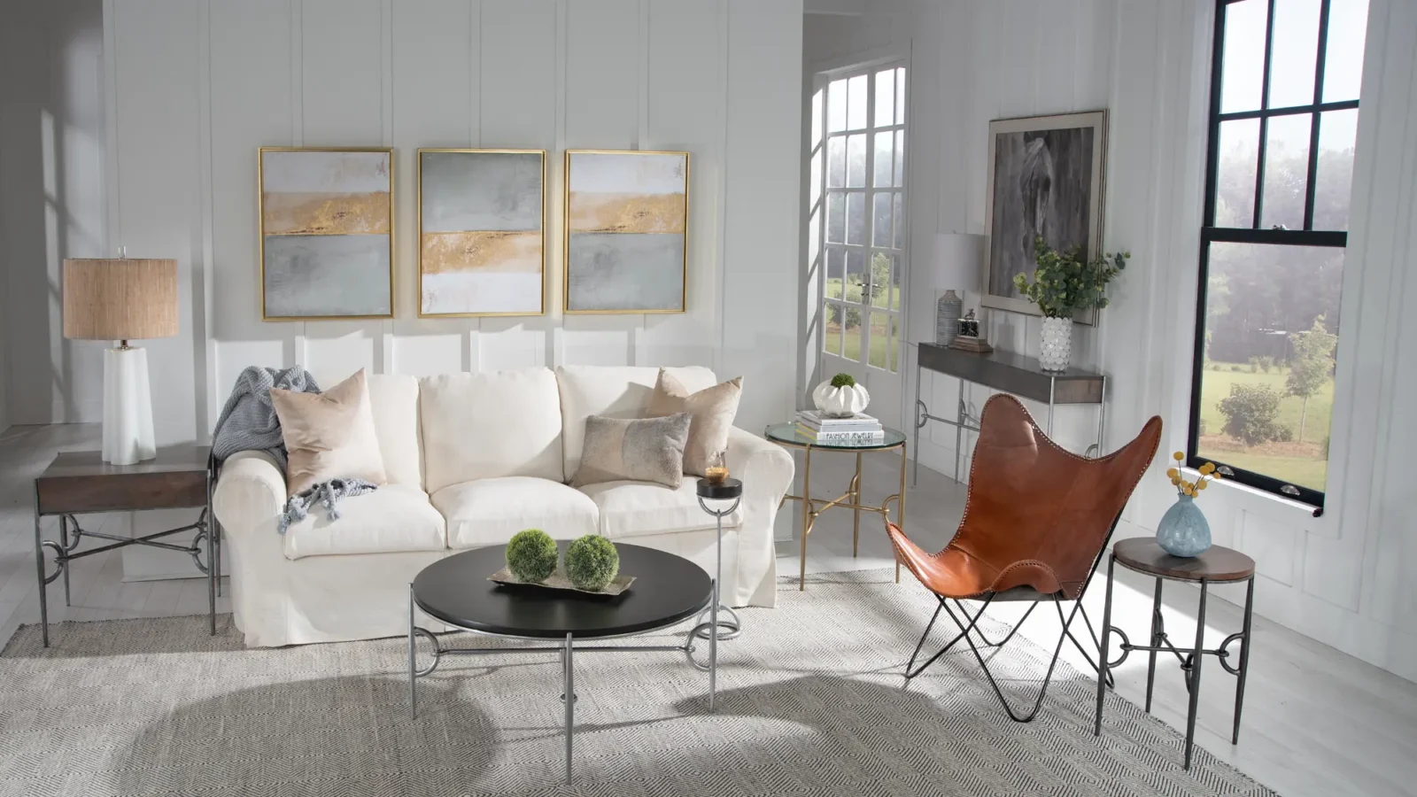 A bright living room featuring furniture pieces from The Paddock Collection, including a black coffee table and various end tables. Three abstract gold and gray paintings hang on the white paneled wall.