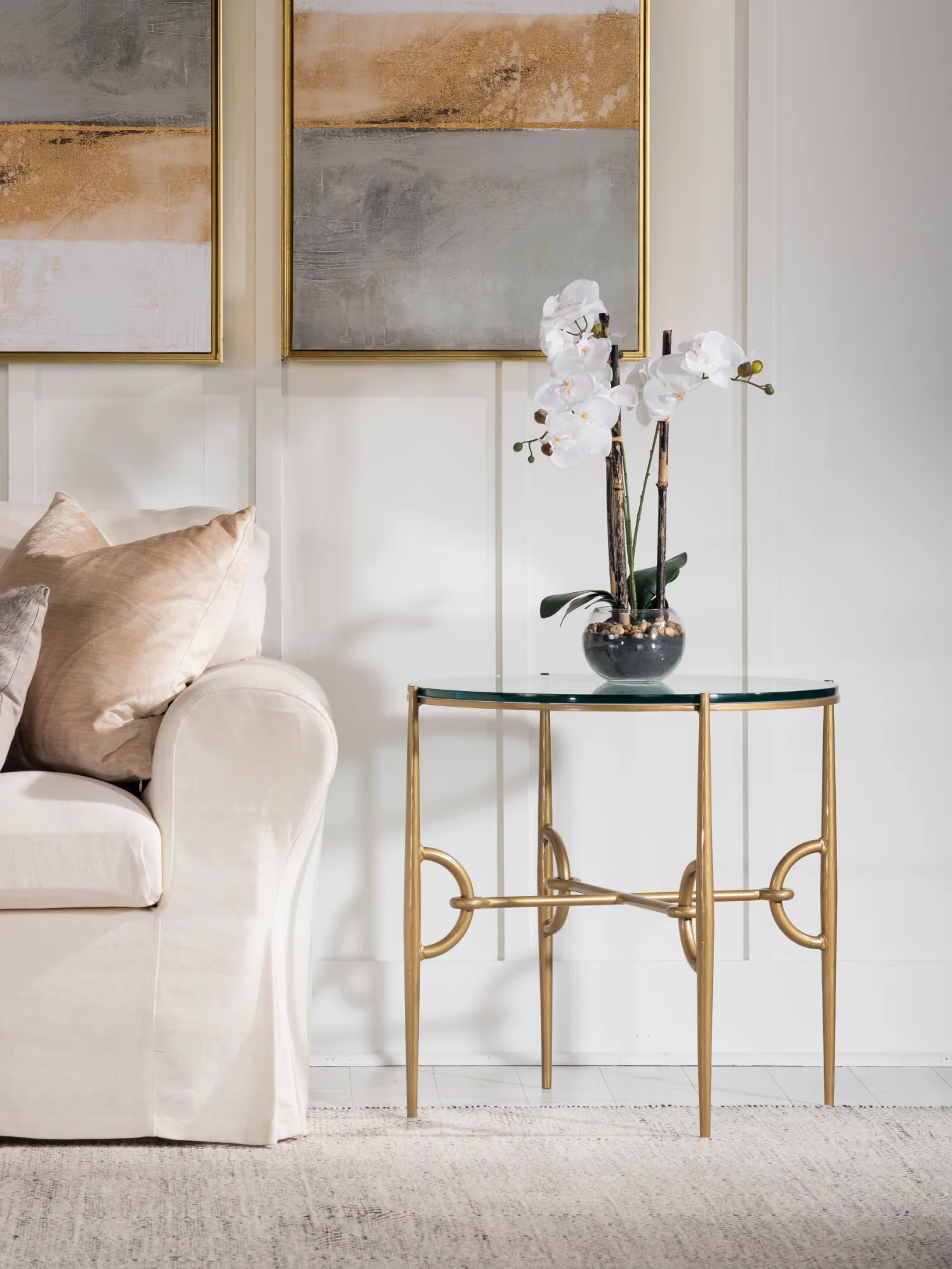 The Paddock Collection round end table, with a glass top and gold base, sits beside a white sofa with tan pillows. A white orchid is displayed on the table.