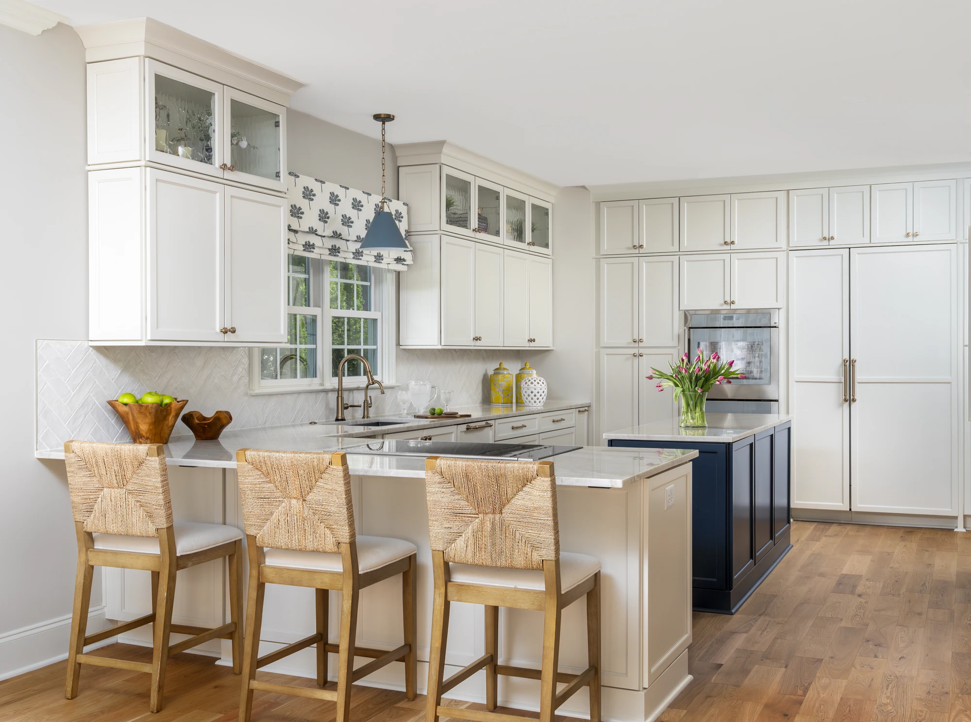 A spacious white kitchen featuring light wood floors and marble surfaces. A peninsula with woven bar stools is contrasted by a small navy island with a floral centerpiece.