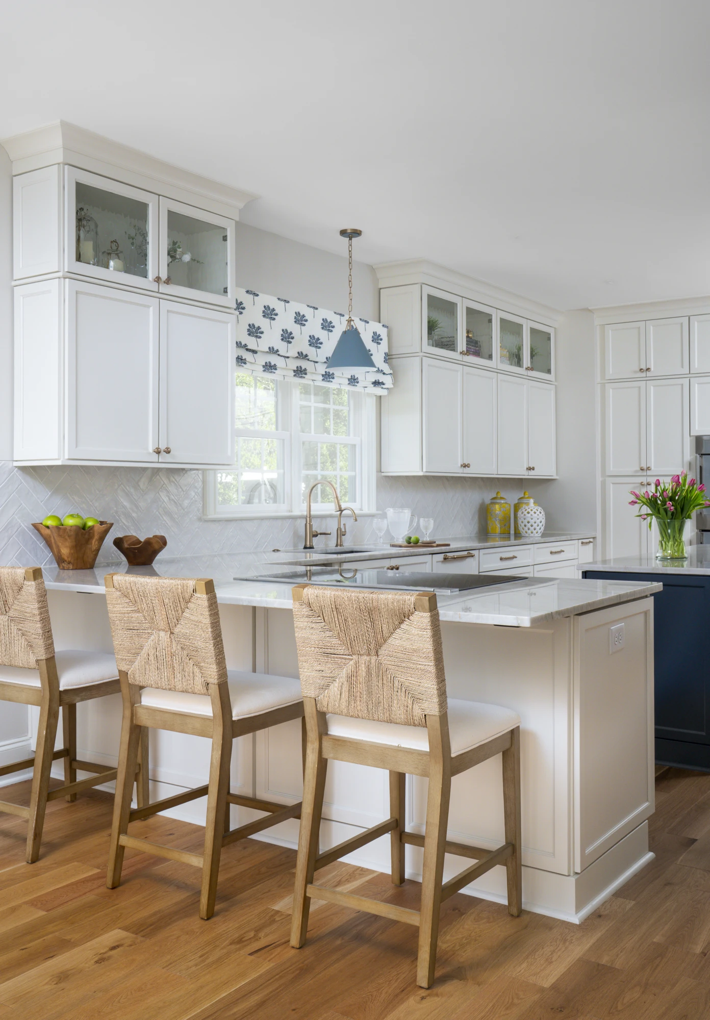A bright, modern kitchen featuring white shaker cabinets, a marble-top island, and light wood flooring. Three woven bar stools line the island.