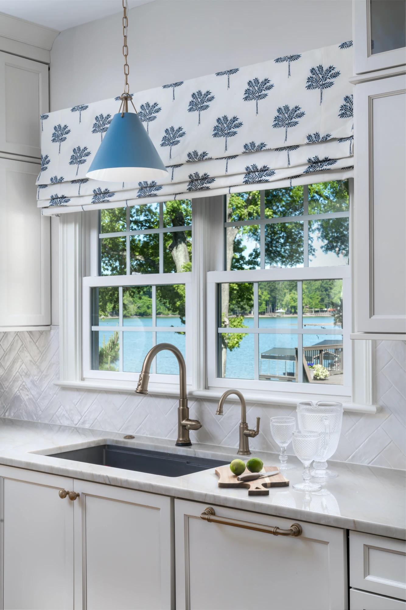 A white kitchen sink area featuring a herringbone backsplash, brass faucets, and a blue conical pendant light. A patterned Roman shade frames a window view of a lake.