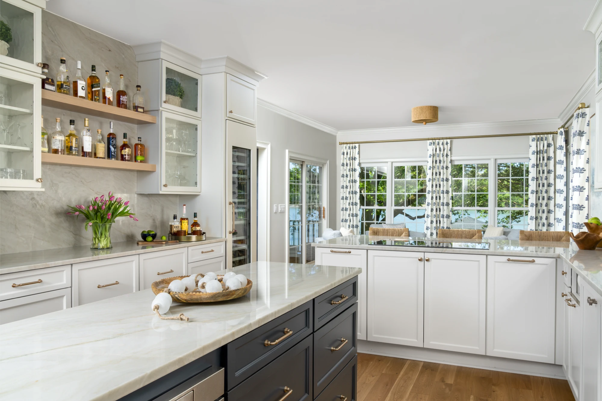 A bright, upscale kitchen featuring white cabinetry, a dark island, and marble countertops. Open shelves display liquor bottles, and large windows show a green, outdoor view.