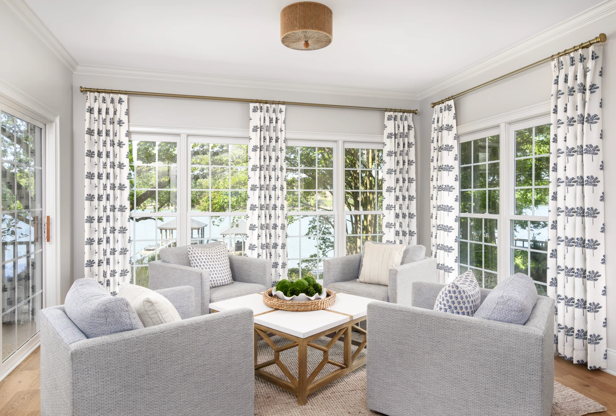 An elegant sitting room features four gray armchairs facing a waterfront view. The space includes a white coffee table and windows dressed with blue botanical-print curtains.