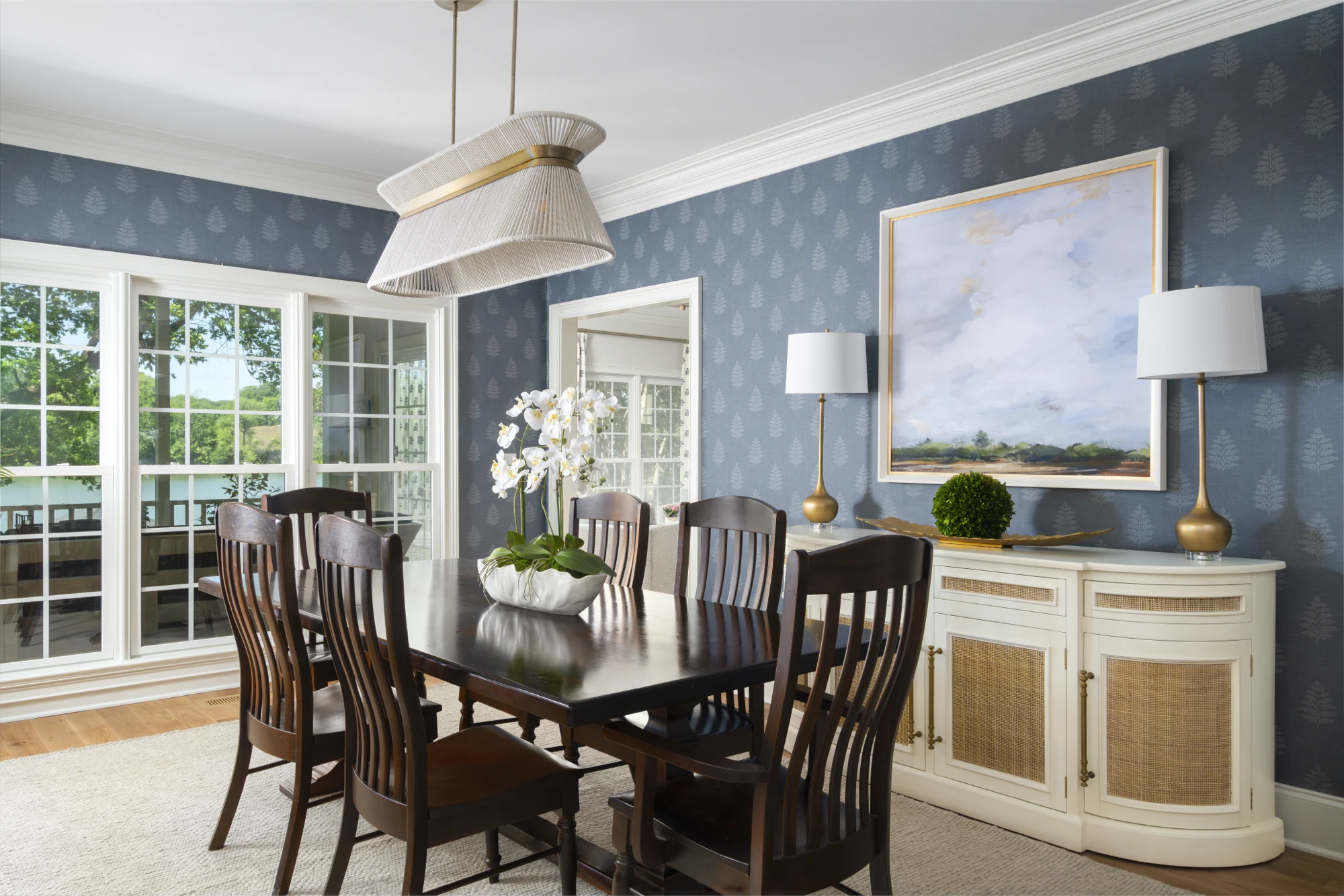 A coastal-inspired dining room featuring blue botanical wallpaper, a dark wood table, and a white rattan sideboard under a large, unique pendant light. Large windows overlook a sunny, green landscape.