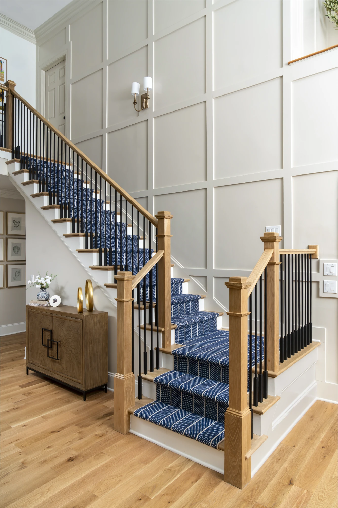 A stylish residential foyer features a staircase with a navy blue and white striped runner, light oak handrails, and black balusters against grid wainscoting.