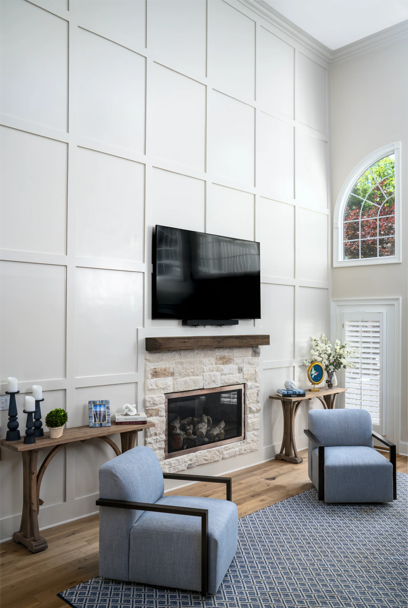 A modern, high-ceiling living room features a tall white wainscoting wall, a stone fireplace, and a mounted TV. Two light blue armchairs sit on a blue patterned rug.