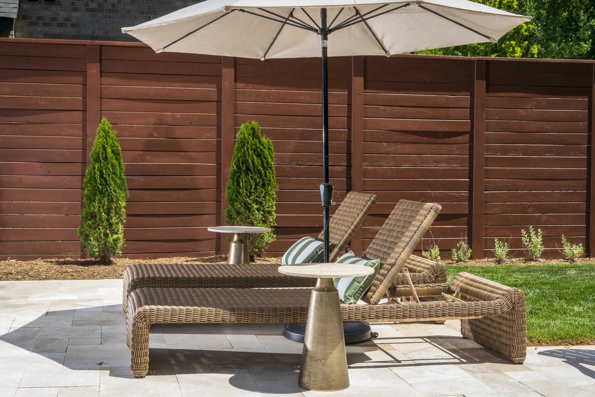 Wicker chaise lounges with striped pillows sit on a light stone patio under a cream umbrella. Two gold side tables are placed near the chairs, against a dark brown wood fence and green lawn.