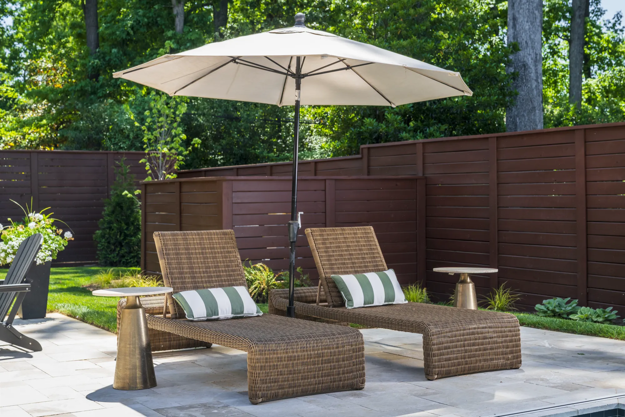 Two woven chaise lounges and small brass side tables are placed on a stone patio, shaded by a cream umbrella against a modern wooden fence.