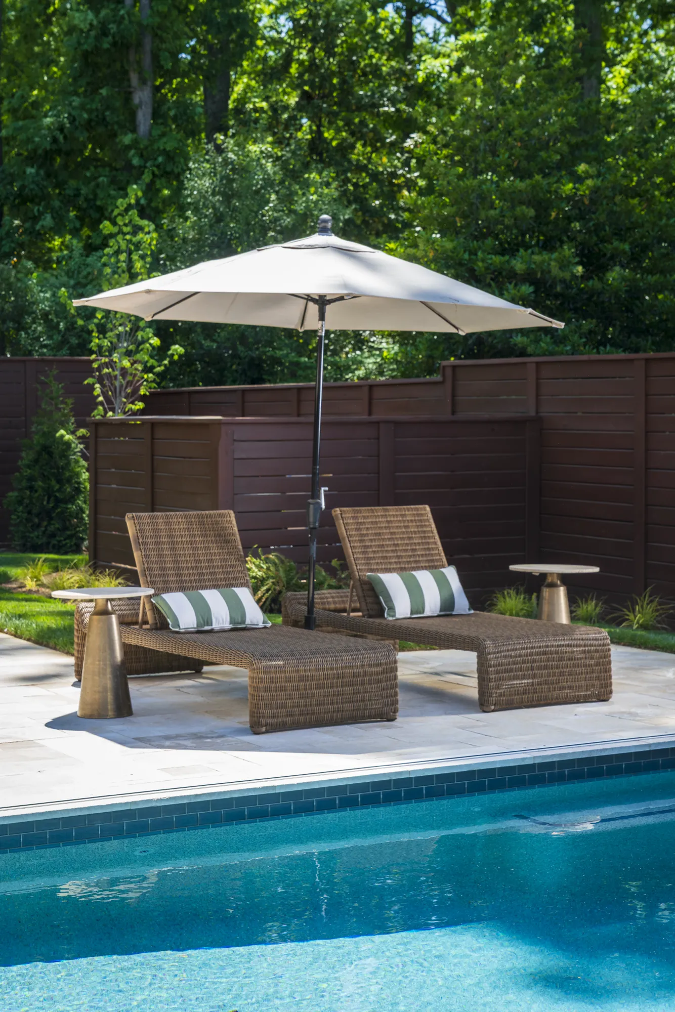 A pair of luxurious woven chaise lounges rest on a sunny poolside patio, shaded by a white umbrella. The chairs feature green and white pillows.