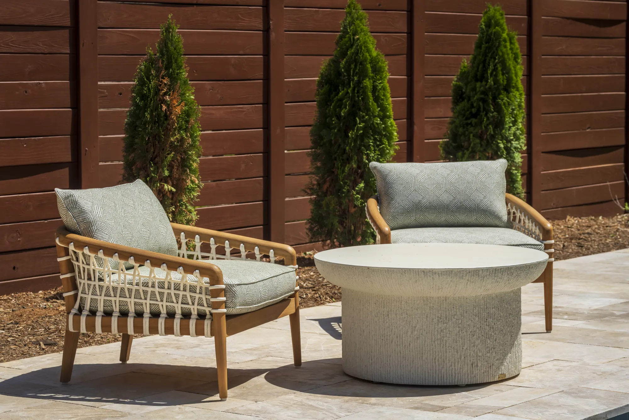 A modern outdoor seating area features two wooden chairs with light green cushions and a round textured table on a stone patio, backed by a dark fence.