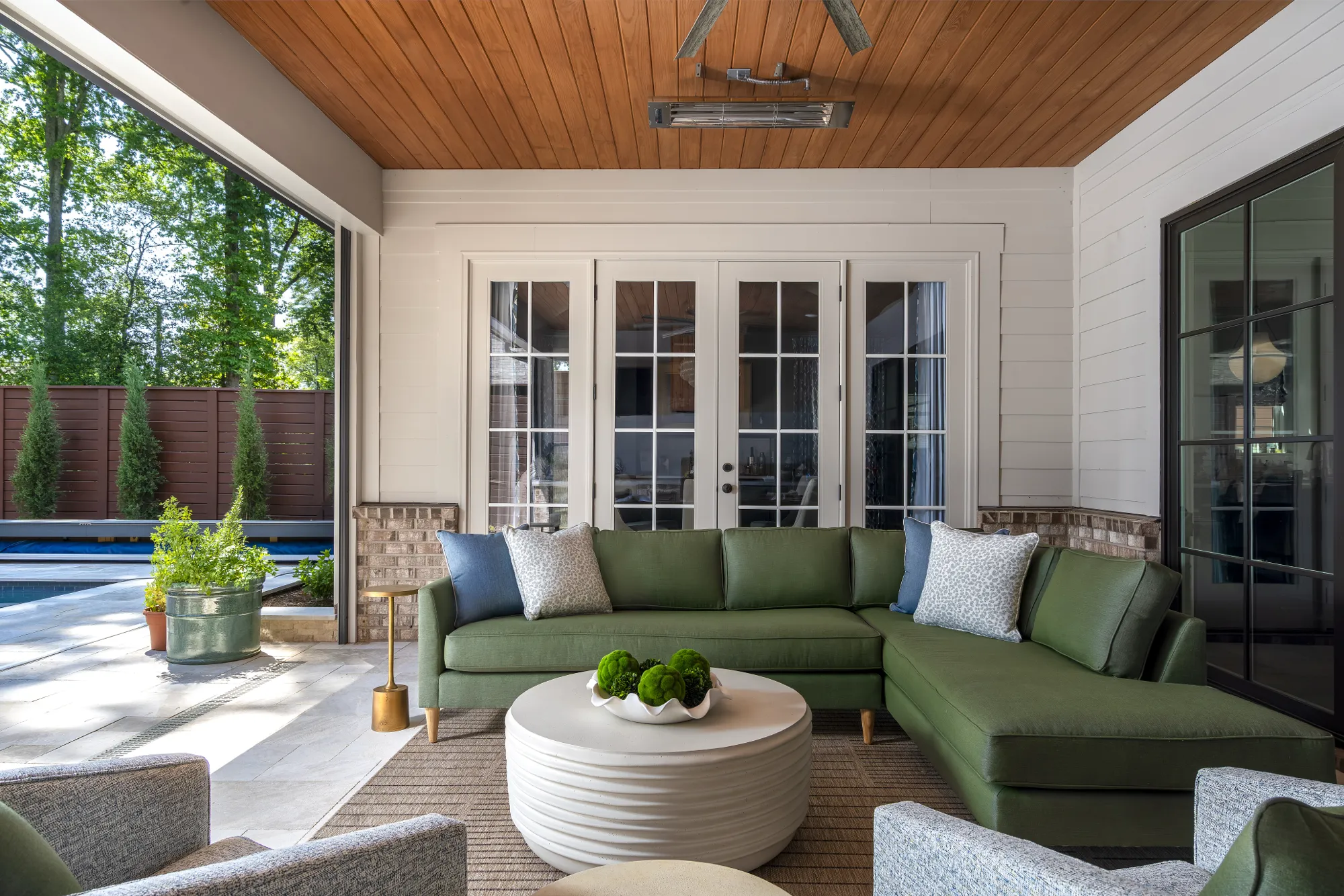An upscale covered patio features an olive green sectional, a round white table, and wood ceiling. The space opens to a bright outdoor pool deck and lush trees.