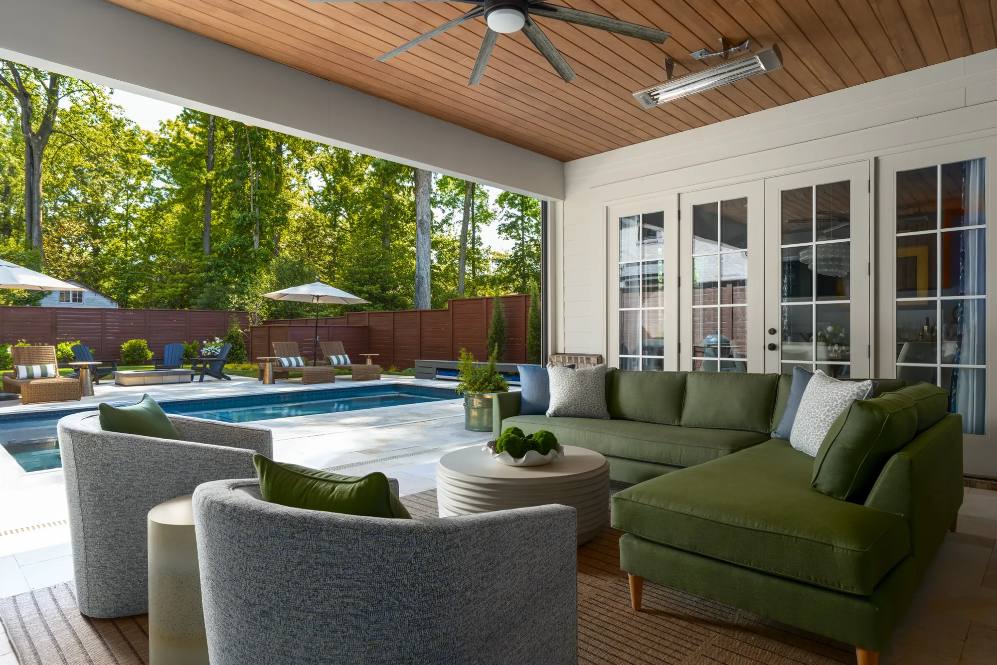 A modern covered patio features an olive green sectional and curved gray chairs, overlooking a swimming pool and a lush, fenced backyard.
