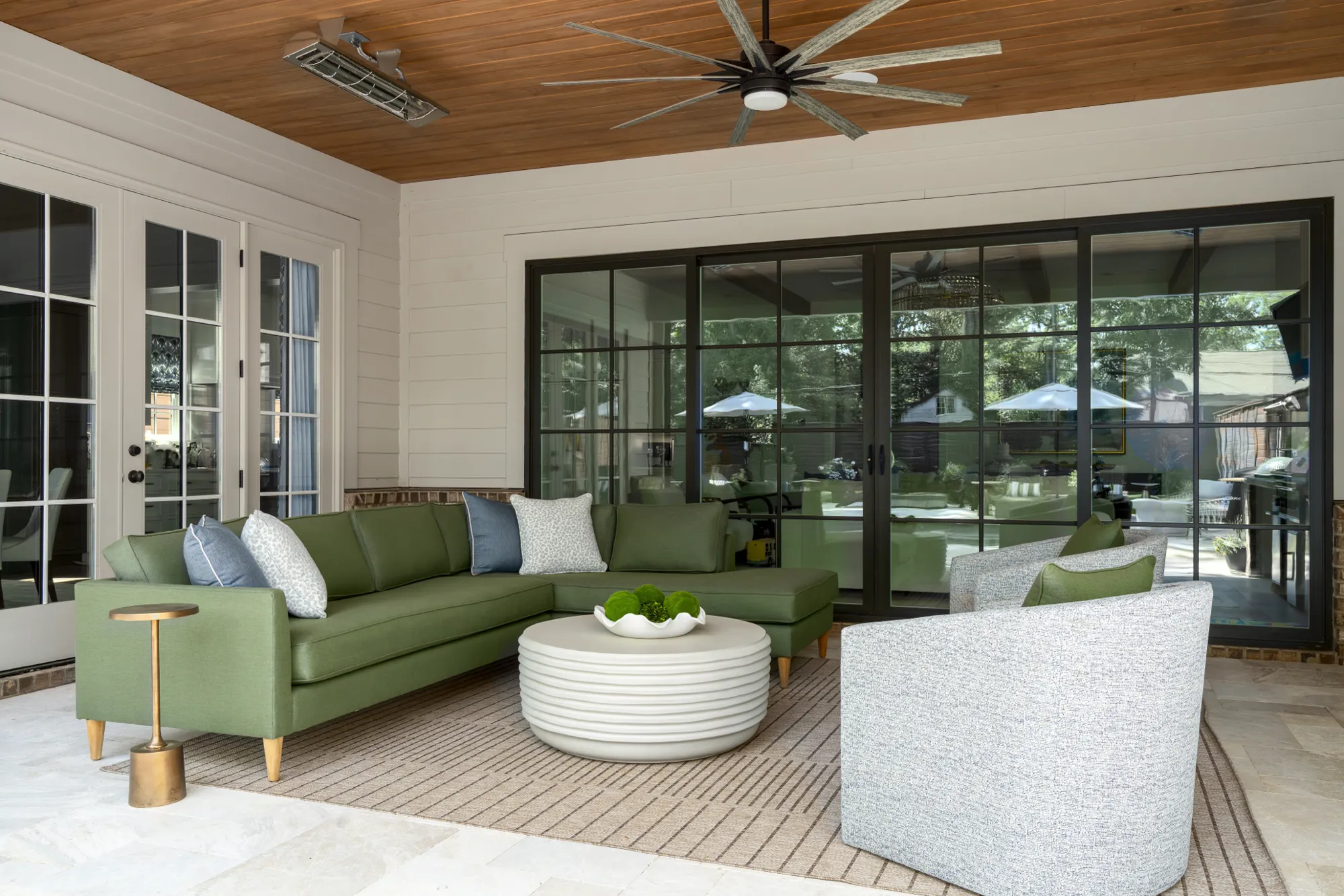 A covered patio with a wood ceiling holds a large olive green sectional, light gray chairs, and a round slatted white coffee table on a neutral rug.
