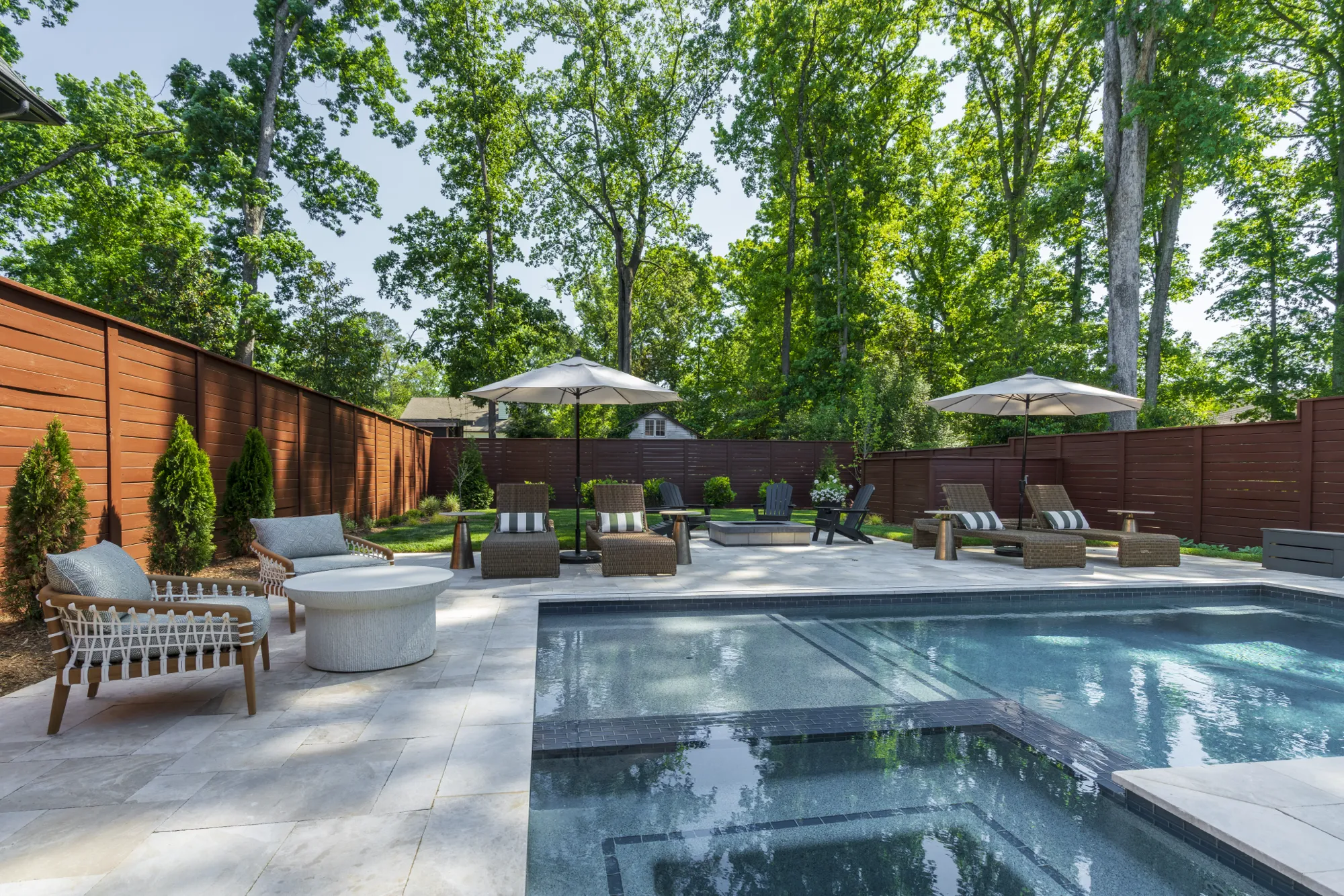 A modern, luxurious backyard swimming pool surrounded by light stone pavers, wicker sun loungers, and casual seating. The private area is enclosed by a dark wooden fence and tall green trees.