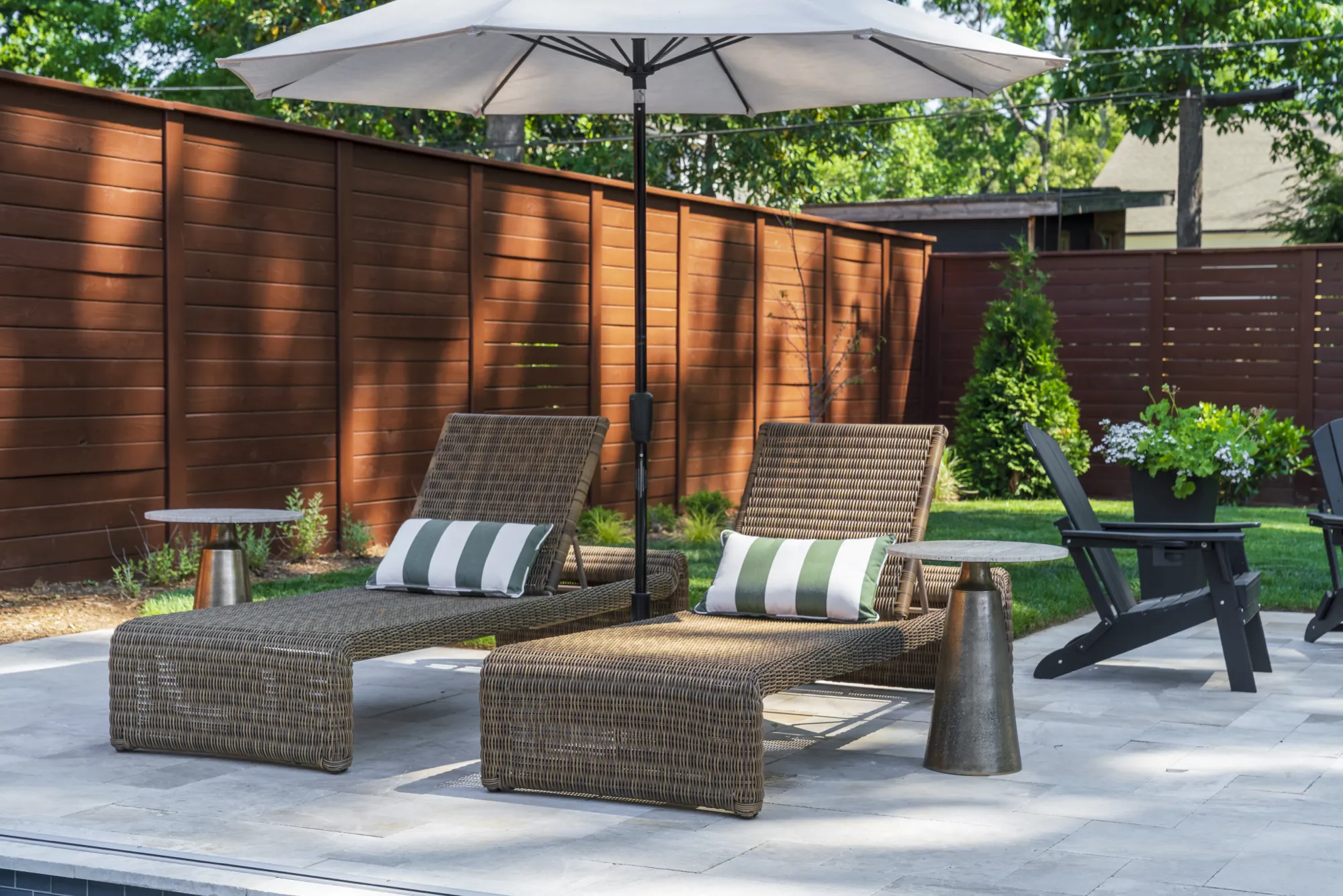 Two woven brown wicker chaises with green and white striped pillows rest on a stone patio under a white umbrella, bordered by a modern dark wood fence.