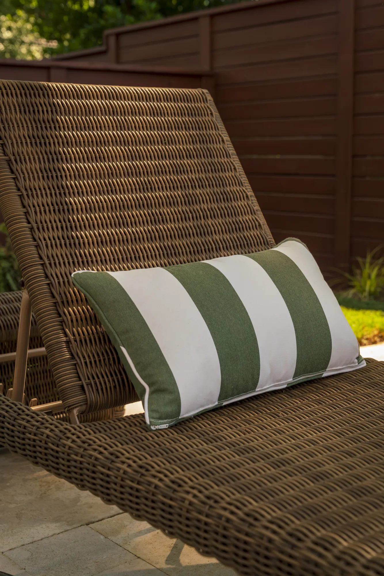 An outdoor brown woven chaise lounge chair rests on a patio, topped with a green and white striped accent pillow. A dark wooden fence is visible in the background.