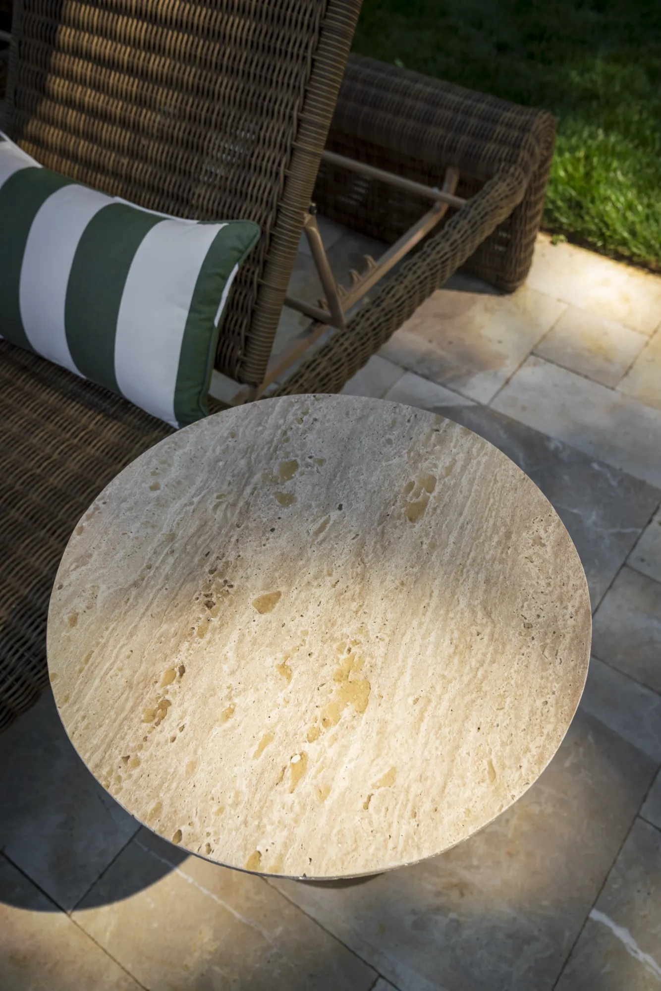 A light-colored, round stone outdoor side table rests on a patio next to a dark woven rattan chair with a striped cushion.