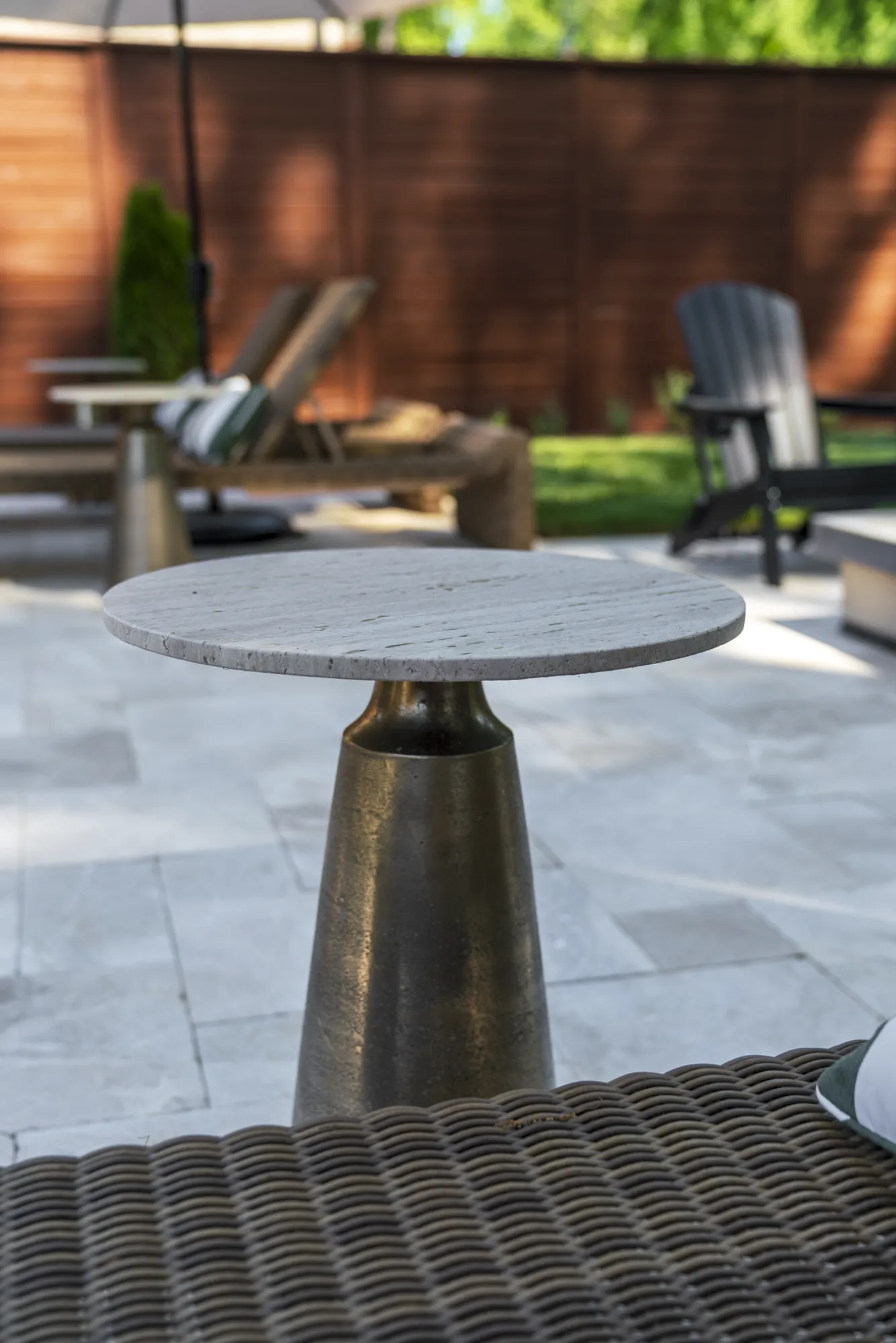 A modern outdoor side table with a light gray stone top and a dark bronze base, set on a tiled patio. Outdoor loungers are blurred in the background.