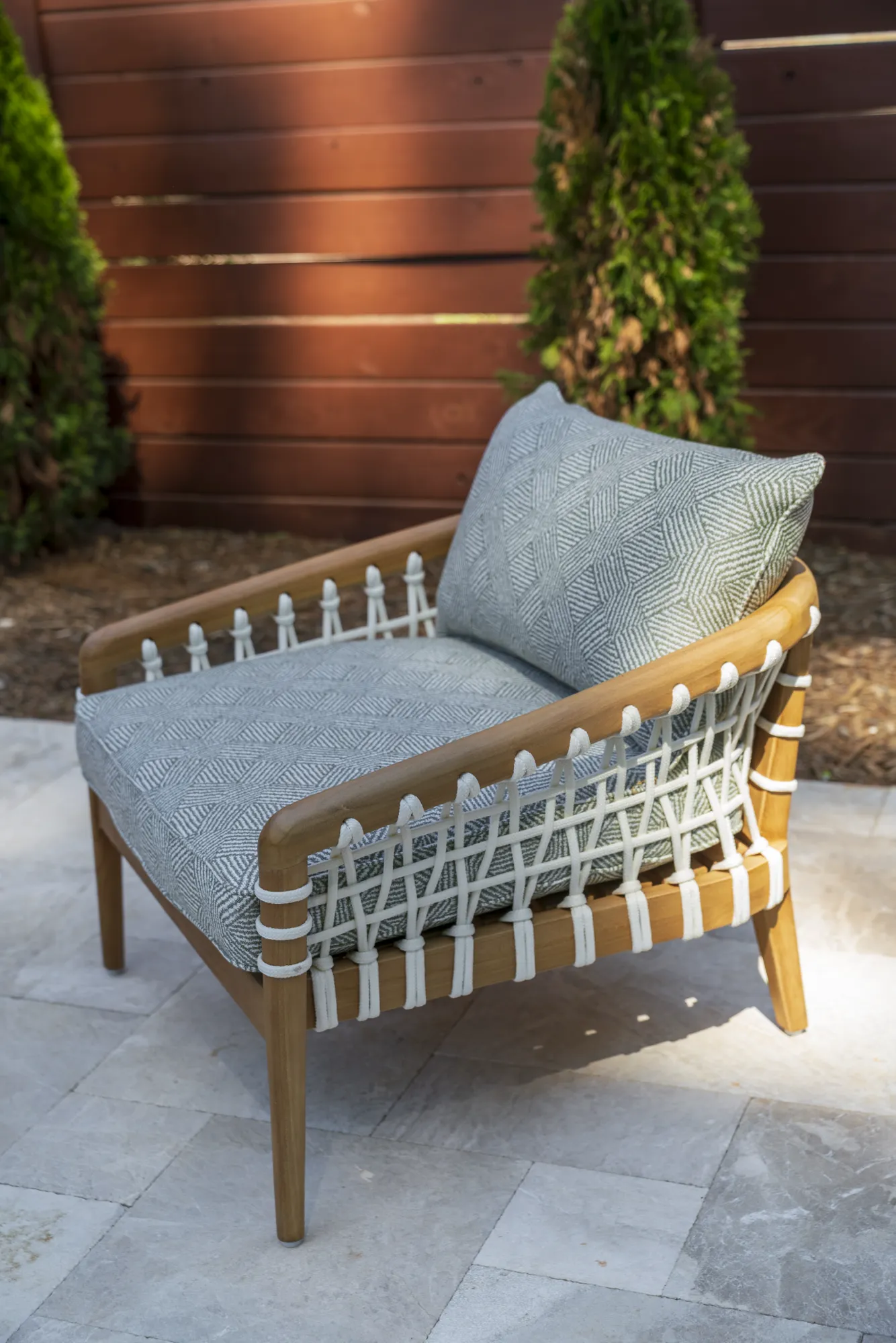 A light wood outdoor lounge chair features white woven rope detailing and thick gray geometric patterned cushions on a stone patio.