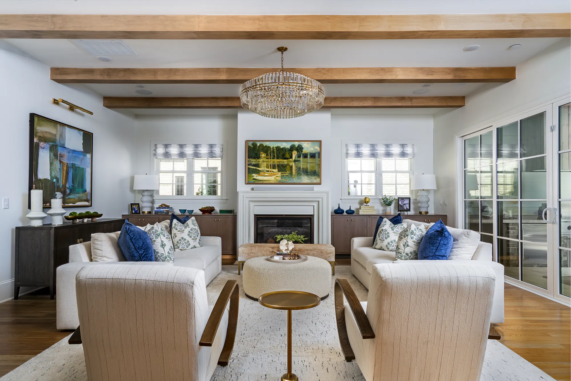 A sophisticated living room with a symmetrical arrangement of white sofas and chairs around an ottoman. The room features exposed wooden ceiling beams, a crystal chandelier, and a central fireplace.