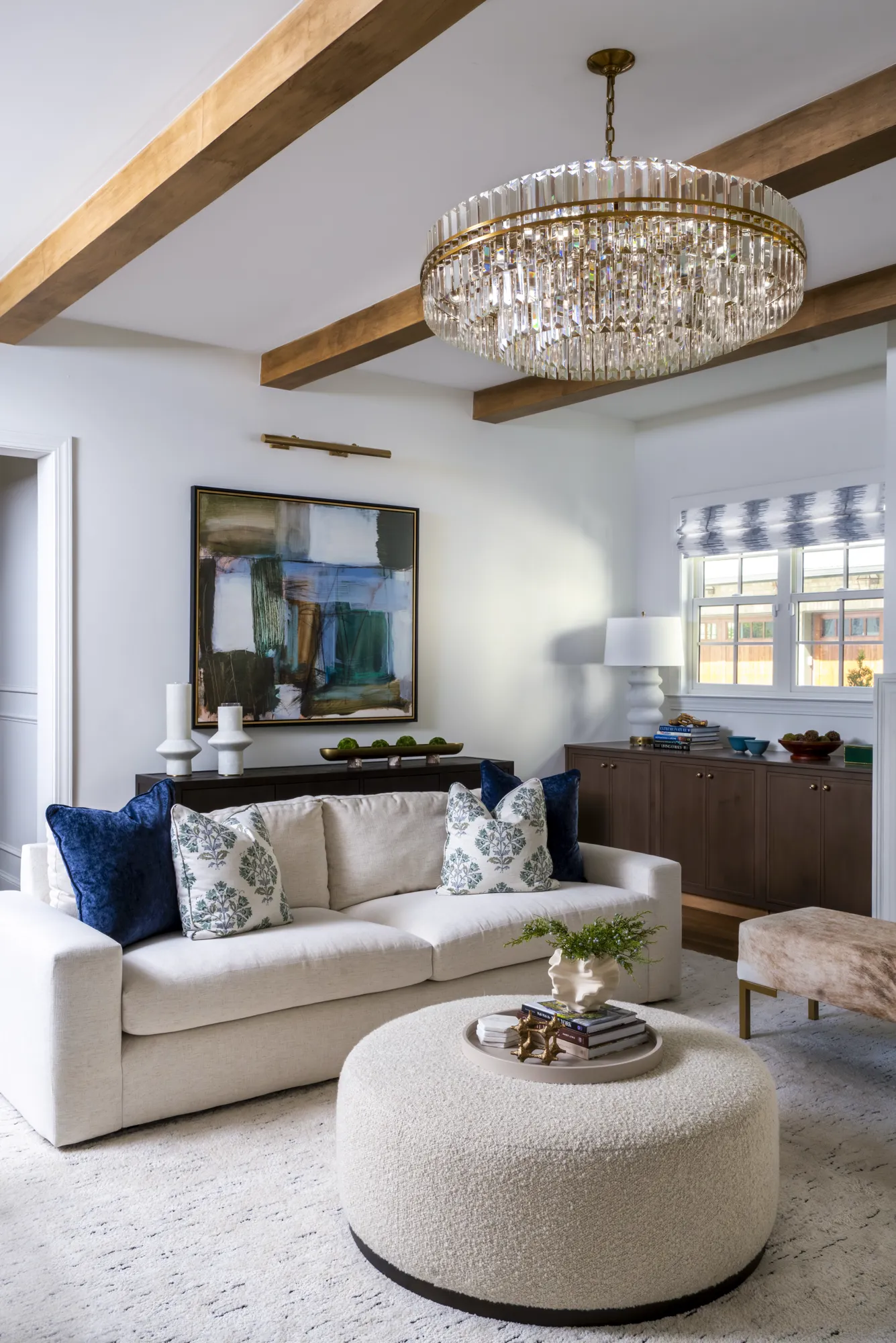 A bright living room with a white sofa and a large abstract blue painting. A crystal chandelier and exposed wood beams draw attention to the ceiling.