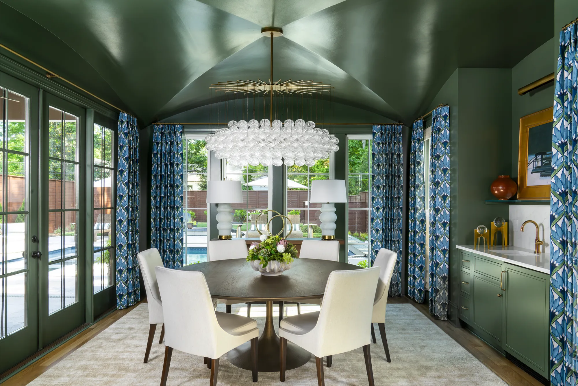 A luxurious dining space with deep green walls and vaulted ceiling. A round table with six white chairs is centered under a large bubble chandelier, next to a small green wet bar.