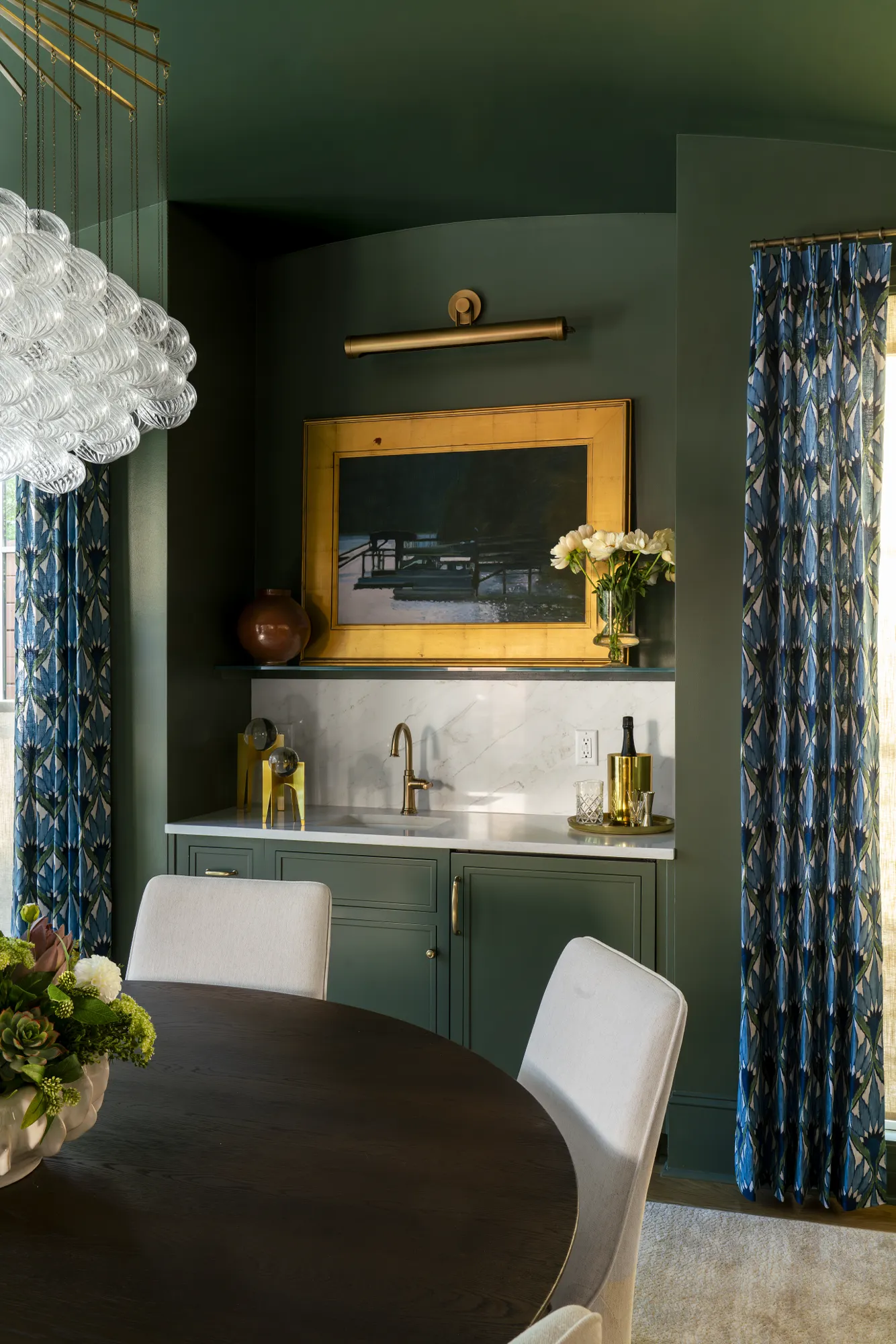A luxurious dining nook features a wet bar with forest green cabinets and marble. The walls are deep green, complemented by patterned blue curtains, a dark wood table, and plush white chairs.