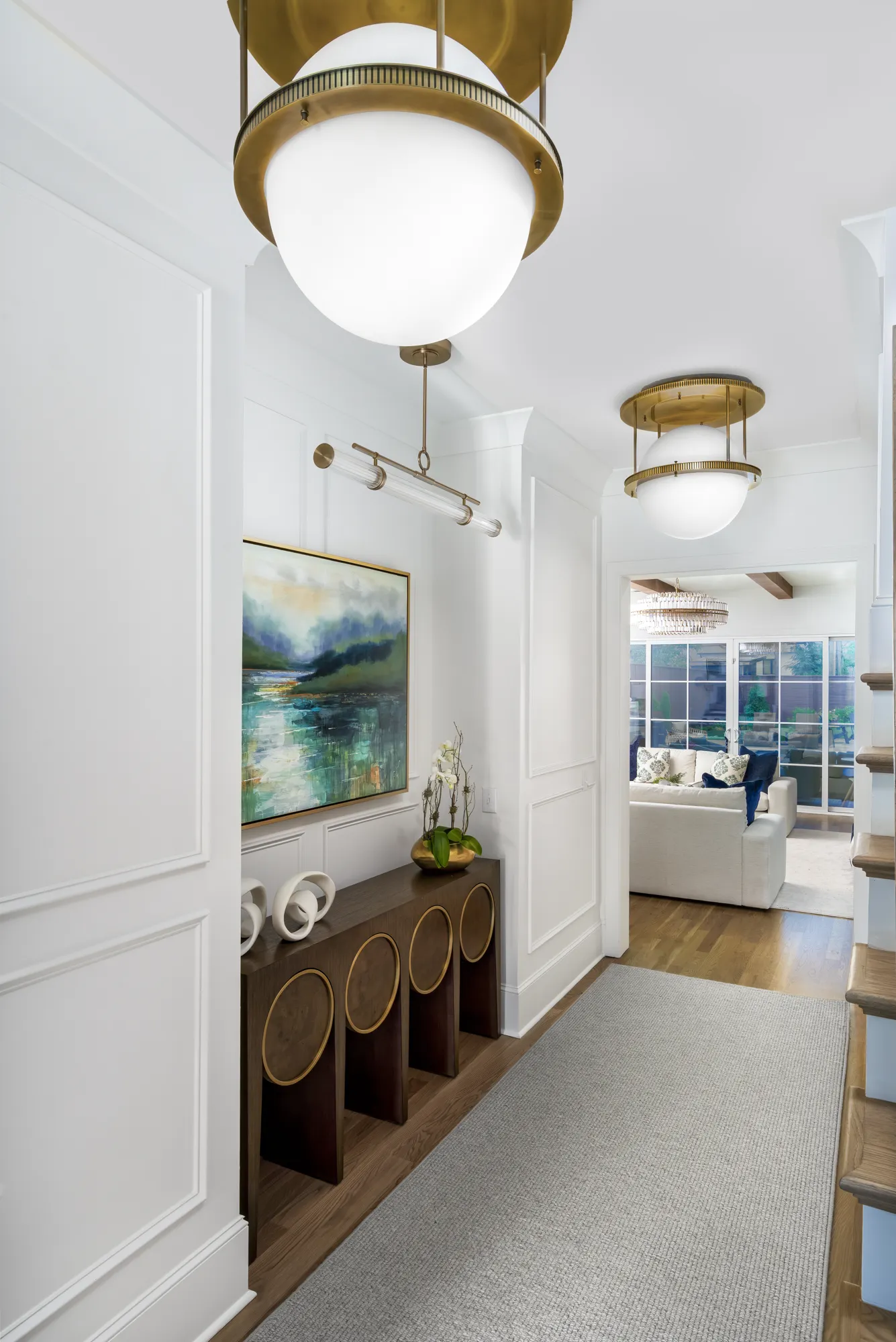 A luxurious white entryway features brass pendant lights, a dark wood console table beneath abstract art, and a long gray runner rug on the hardwood floor.