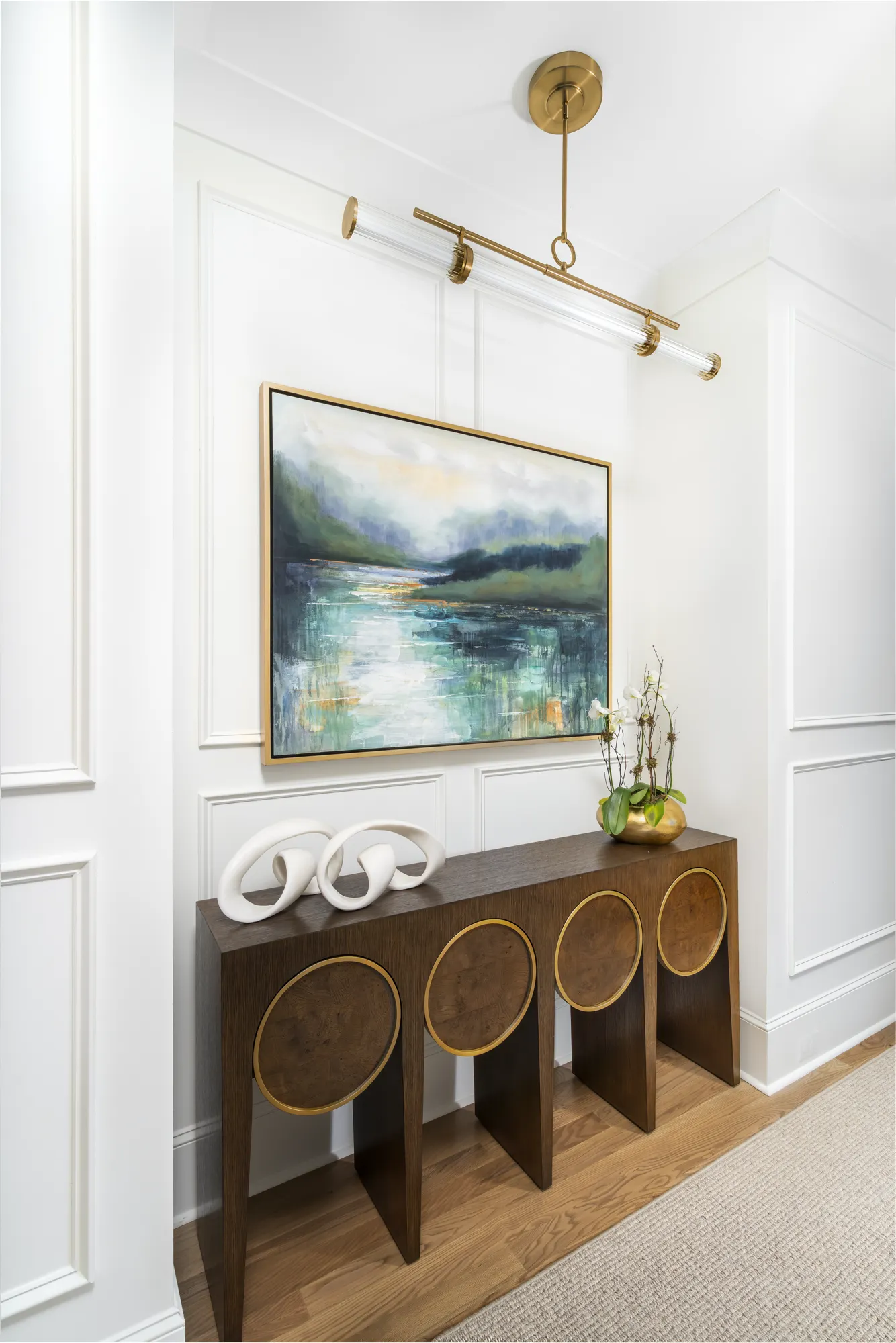 A dark wood console table with brass accents sits against a white wainscoted wall. Above it is an abstract landscape painting, flanked by modern white sculptures and a gold orchid planter.