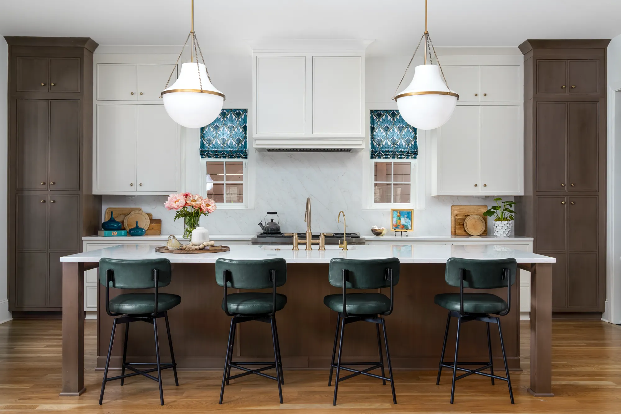 A modern residential kitchen showing a large island with four green stools. The design includes two-toned brown and white cabinets, a marble backsplash, and brass fixtures.