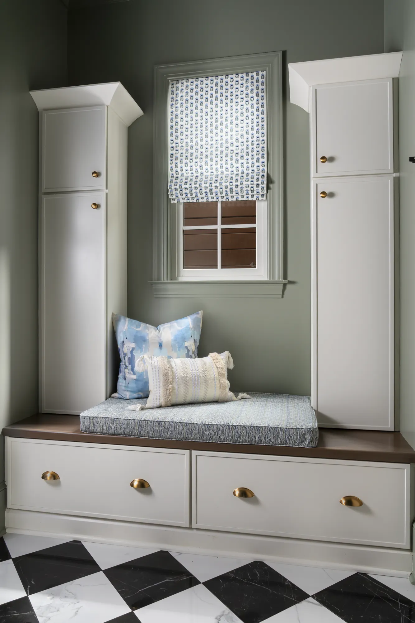 A built-in bench with storage drawers and flanking cabinets features a blue cushion and patterned pillows against sage green walls. The floor is black and white checkerboard tile.