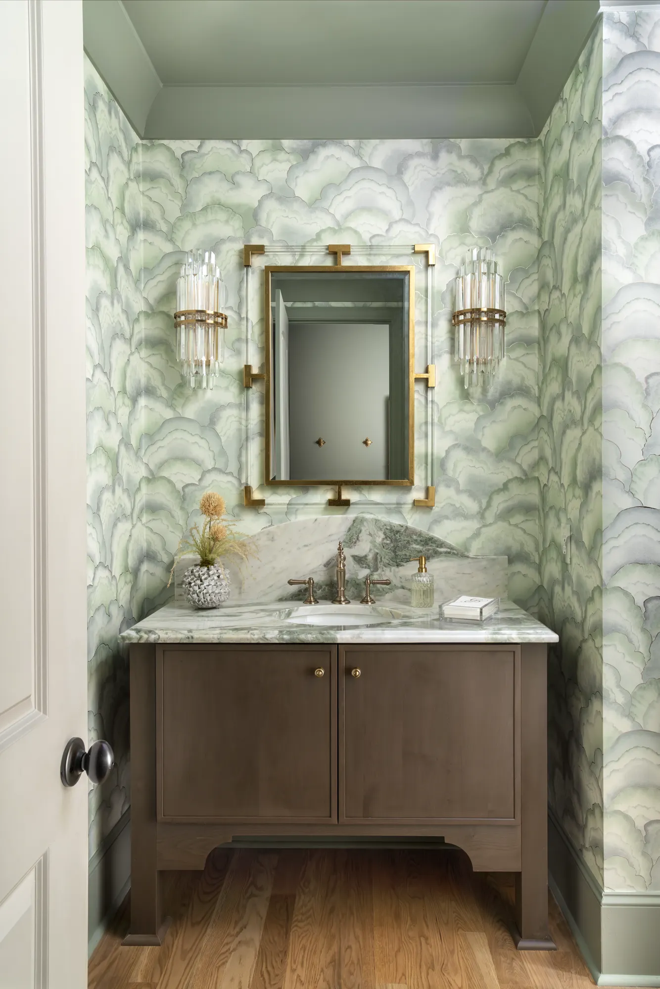 A stylish powder room with green cloud wallpaper, a dark vanity, and a green marble counter centered under a gold mirror and crystal sconces.