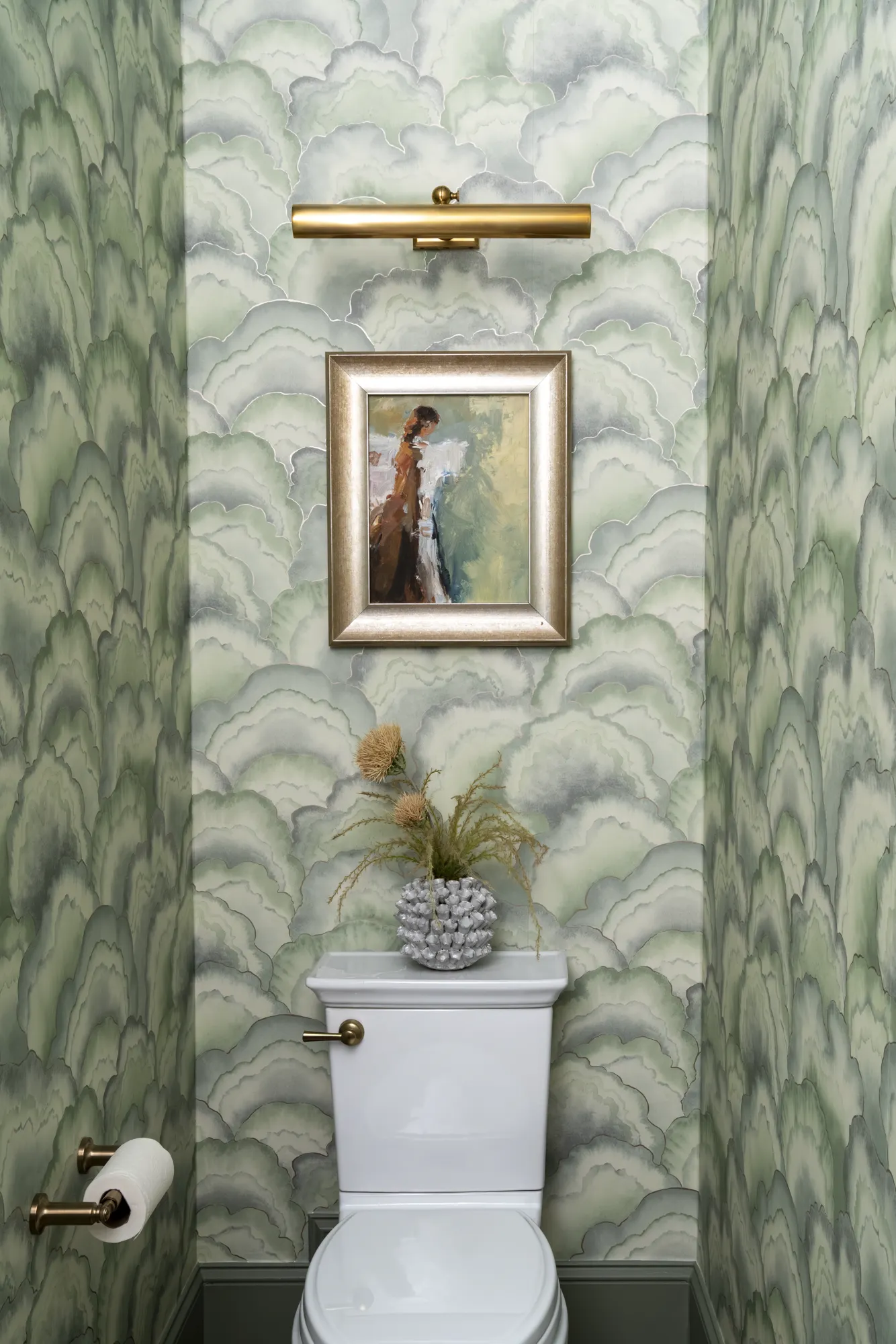 A powder room features striking green scallop wallpaper, brass accents, and a white toilet. A framed painting hangs above the tank.