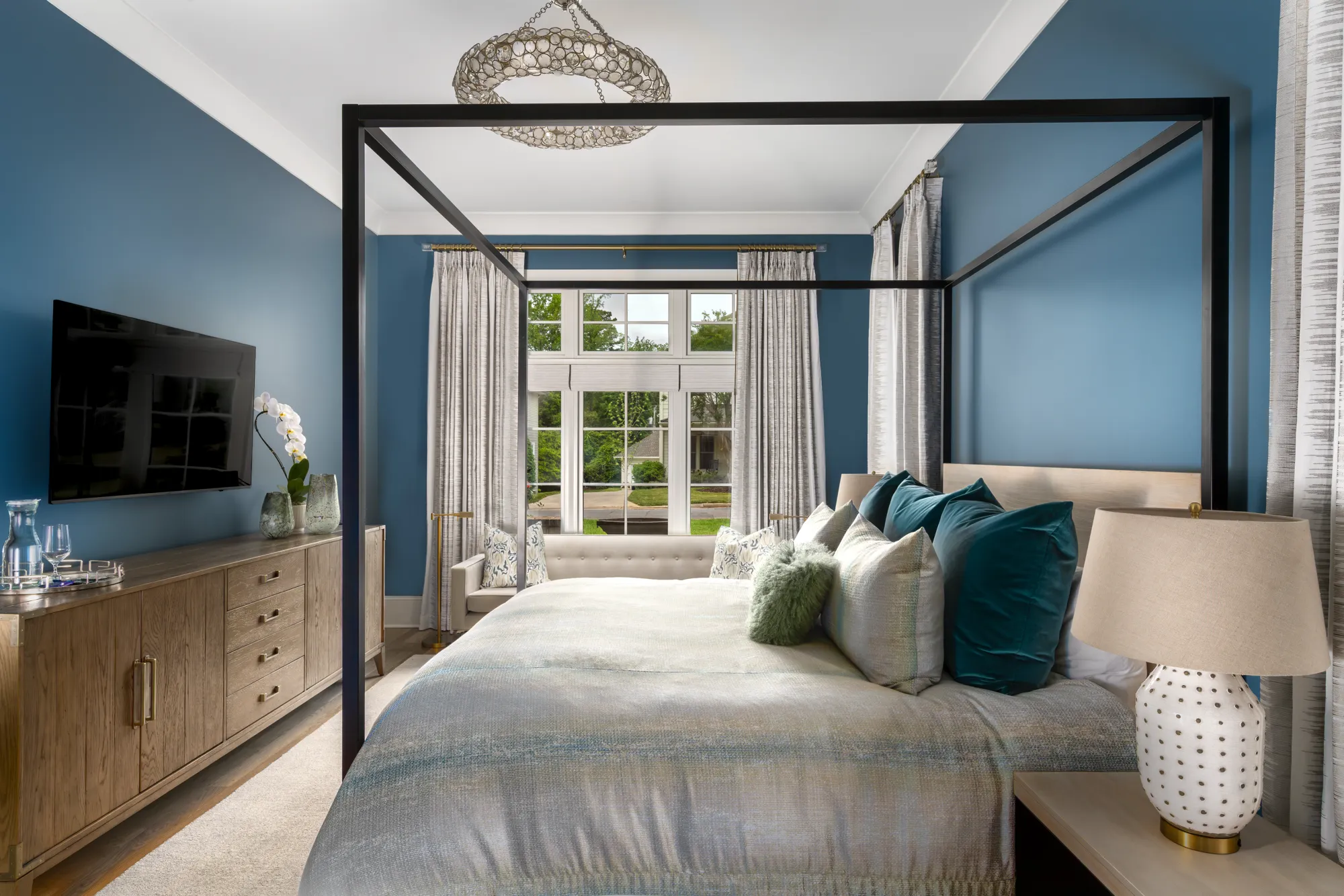 A stylish master bedroom with deep blue walls and a black four-poster bed covered in white and teal bedding. A long wooden console sits beneath a television.
