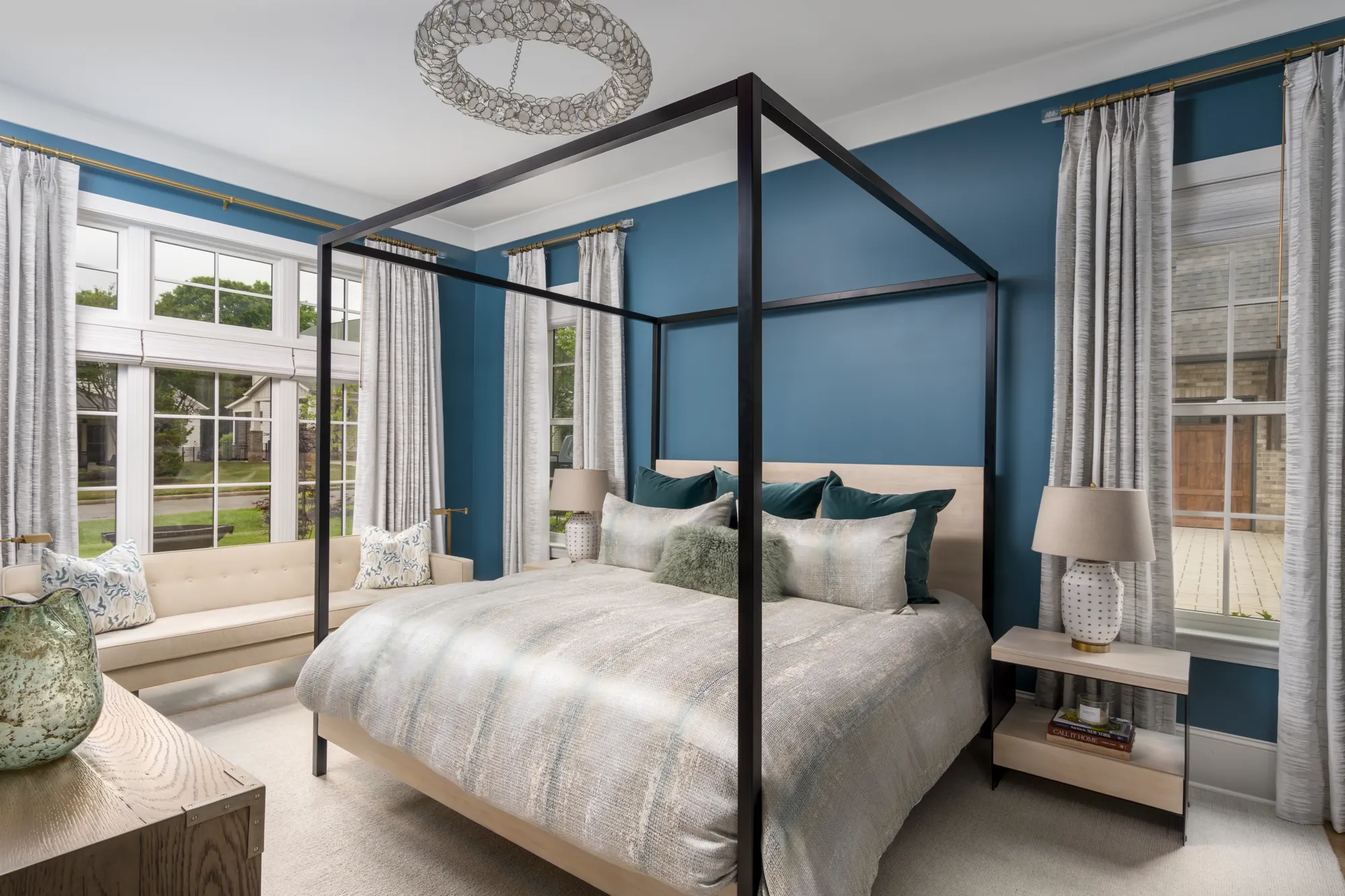A sophisticated bedroom featuring a dark canopy bed, teal accent wall, and luxurious bedding. Large windows frame a beige seating bench and a crystal chandelier hangs overhead.