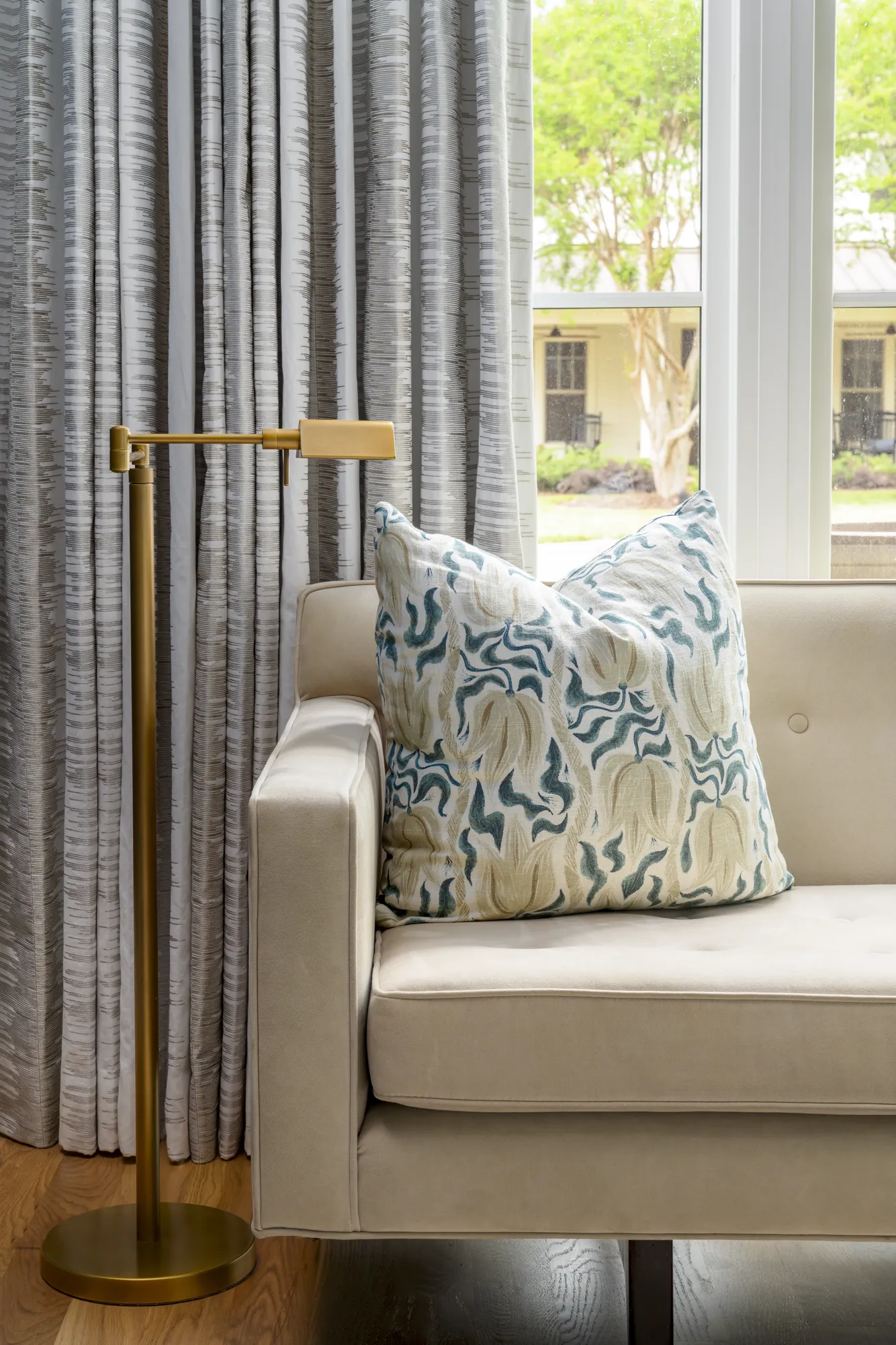 The arm of a cream sofa with a large teal and beige patterned pillow. A gold floor lamp and textured grey drapes frame a bright window.