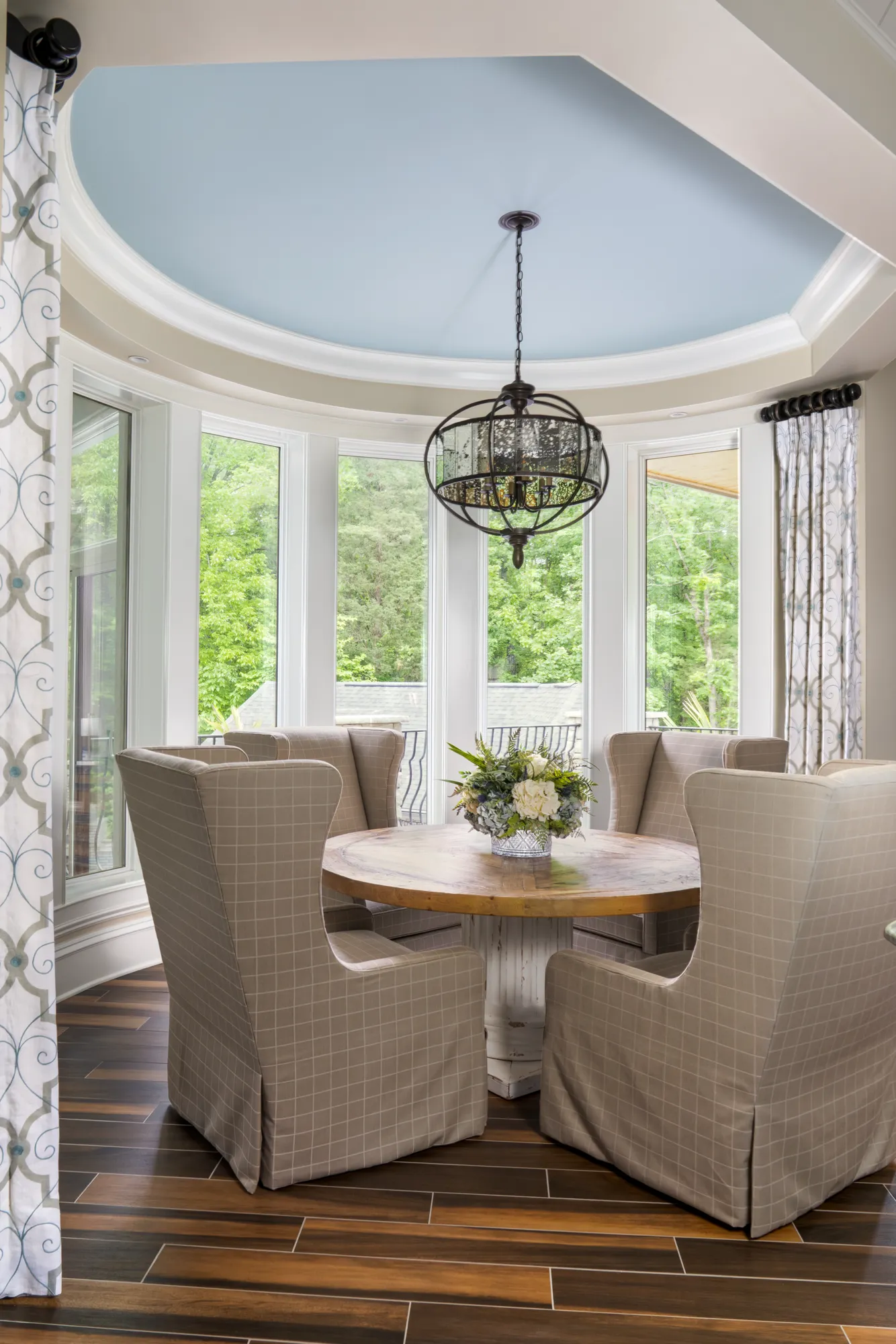 An elegant breakfast nook with a round wood table and beige chairs in a curved bay window alcove. A light blue ceiling and dark metal chandelier complete the design overlooking nature.
