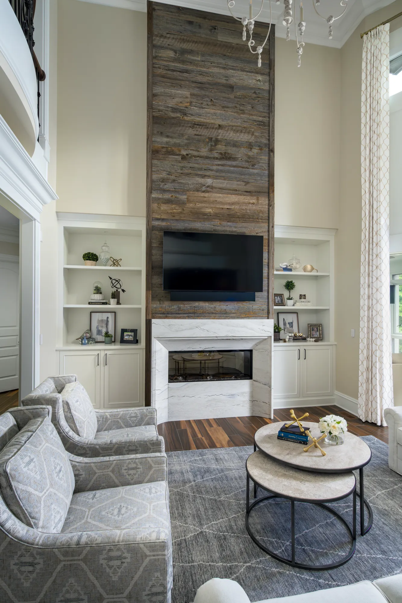 A transitional living room featuring a tall fireplace wall covered in reclaimed wood paneling, flanked by white built-in shelving. Upholstered armchairs sit near nesting coffee tables on a gray rug.