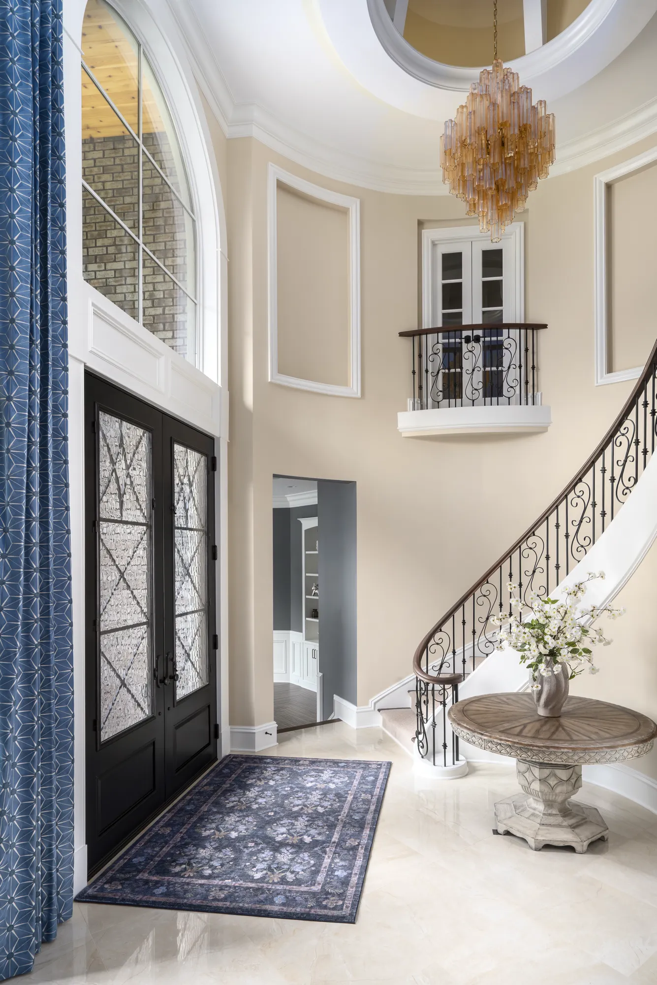 A soaring two-story foyer featuring an ornate crystal chandelier, a curved staircase with ironwork, and black double entry doors set on light marble flooring.