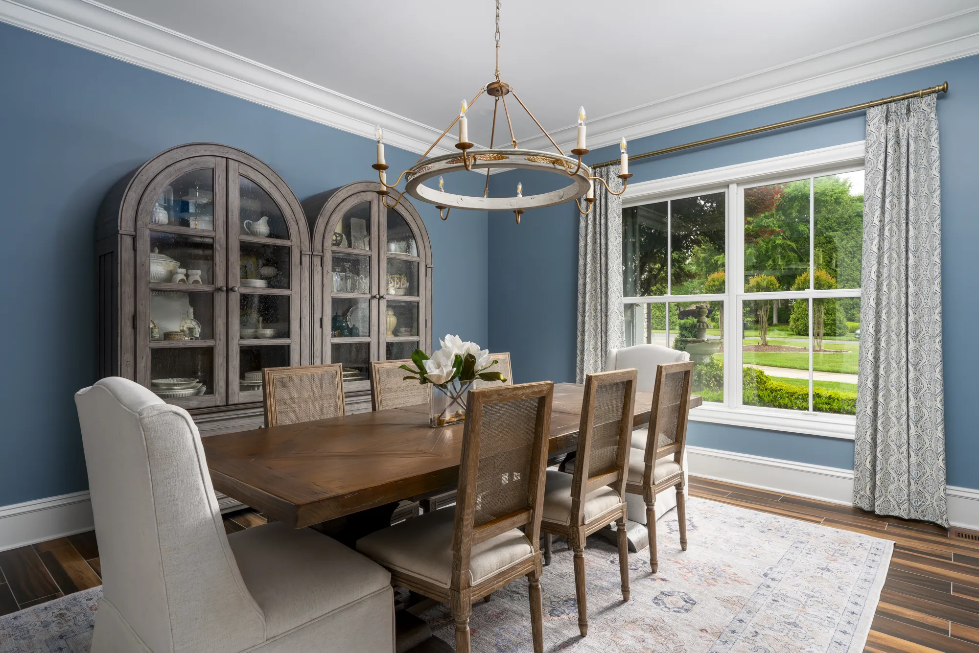 A formal dining room with slate blue walls features a wood table, upholstered and cane-backed chairs, and a round distressed chandelier. Two curio cabinets display white dishware.