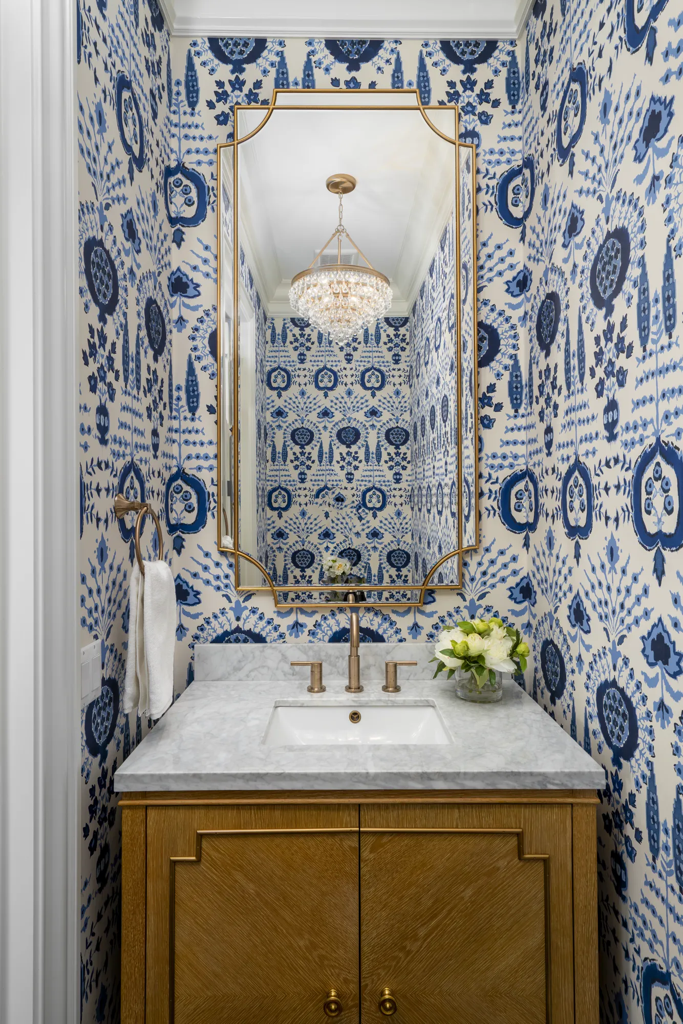 A bright powder room featuring bold blue and white patterned wallpaper. A brass-framed mirror hangs over a wood vanity with a marble counter.