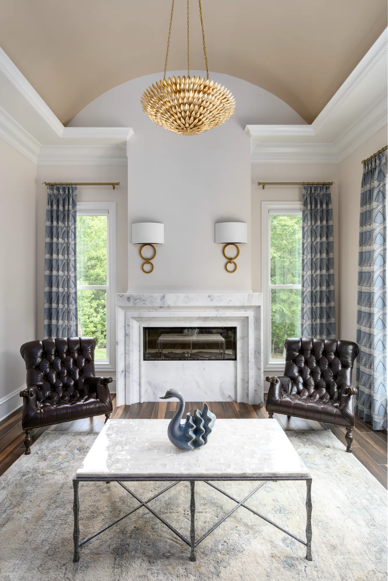 A formal seating area with a white marble fireplace flanked by two dark leather tufted chairs. A unique gold chandelier hangs above a white coffee table featuring a blue swan sculpture.