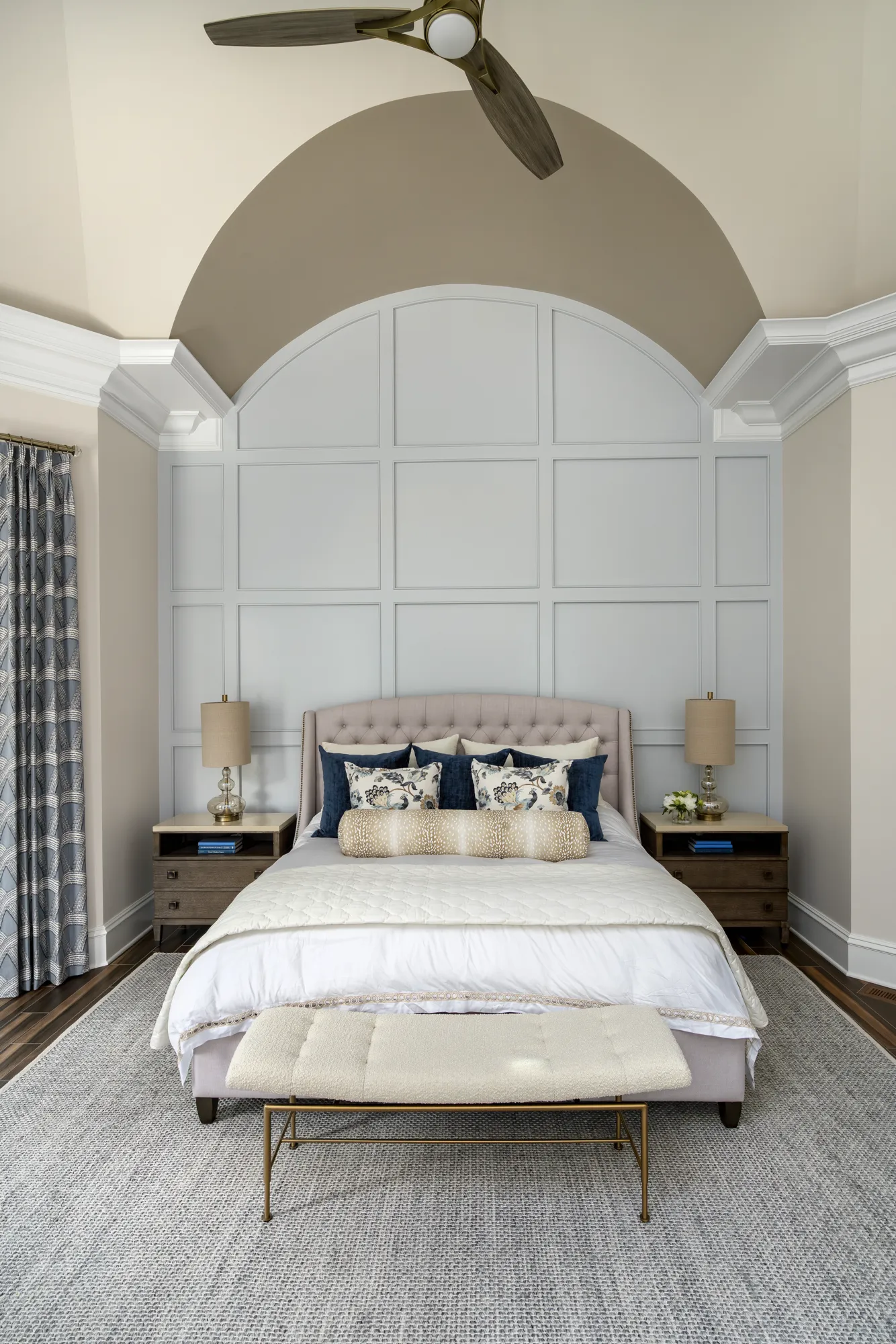 A transitional master bedroom with a tufted bed centered against a light gray paneled accent wall in an arched ceiling alcove. The bed is flanked by dark wood nightstands.