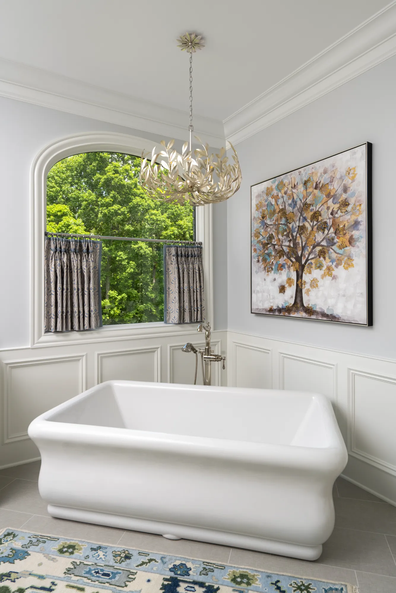 A white freestanding bathtub is centered beneath an arched window looking out to green trees. The room features wainscoting, a leaf chandelier, and an abstract tree painting.